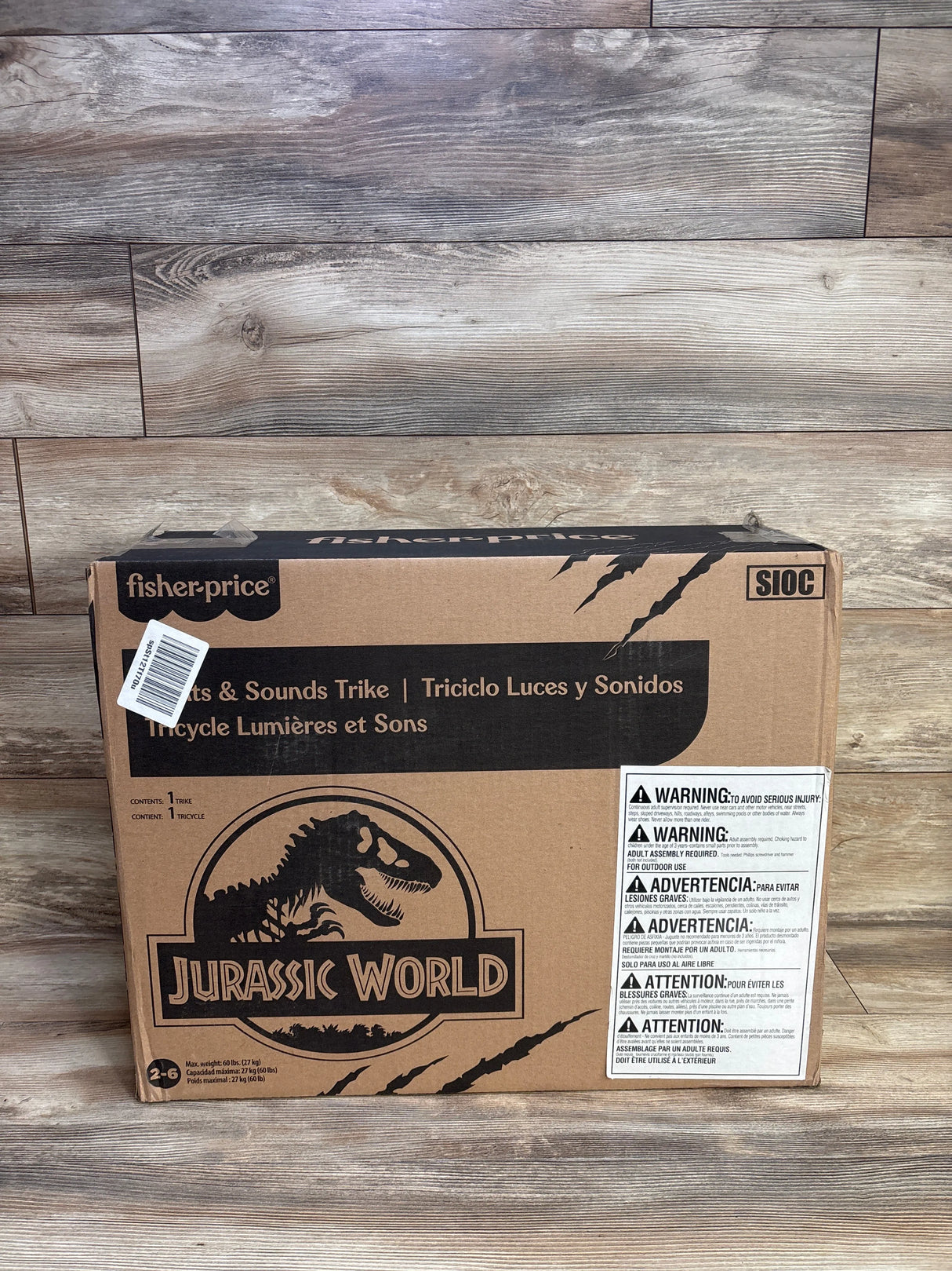 Front view of Fisher-Price Velociraptor tricycle box showing Jurassic World logo and warning label on wood backdrop
