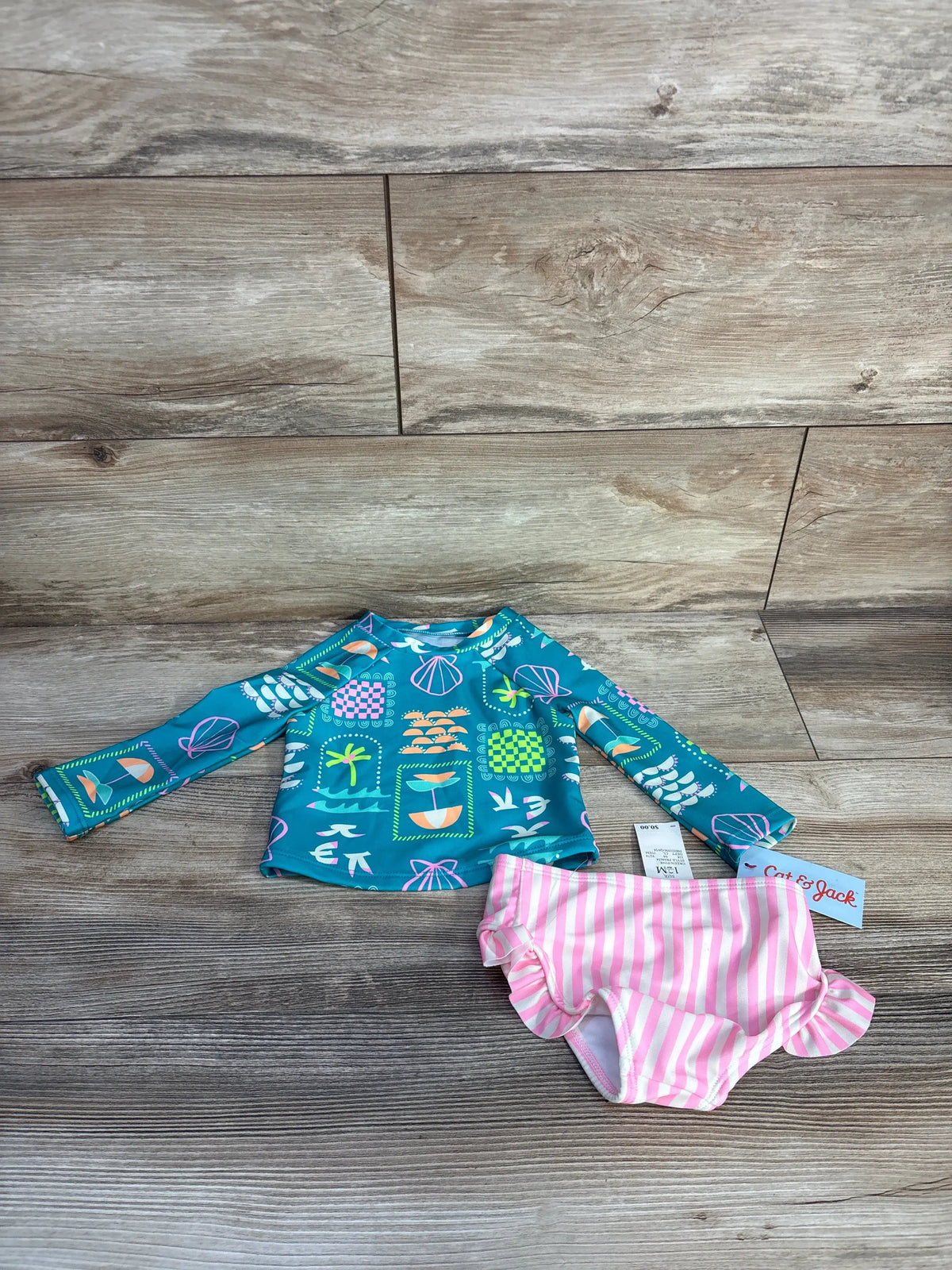 Flat lay of baby rashguard tankini showcasing seafoam top with boat print and pink striped ruffle bottoms on wood background