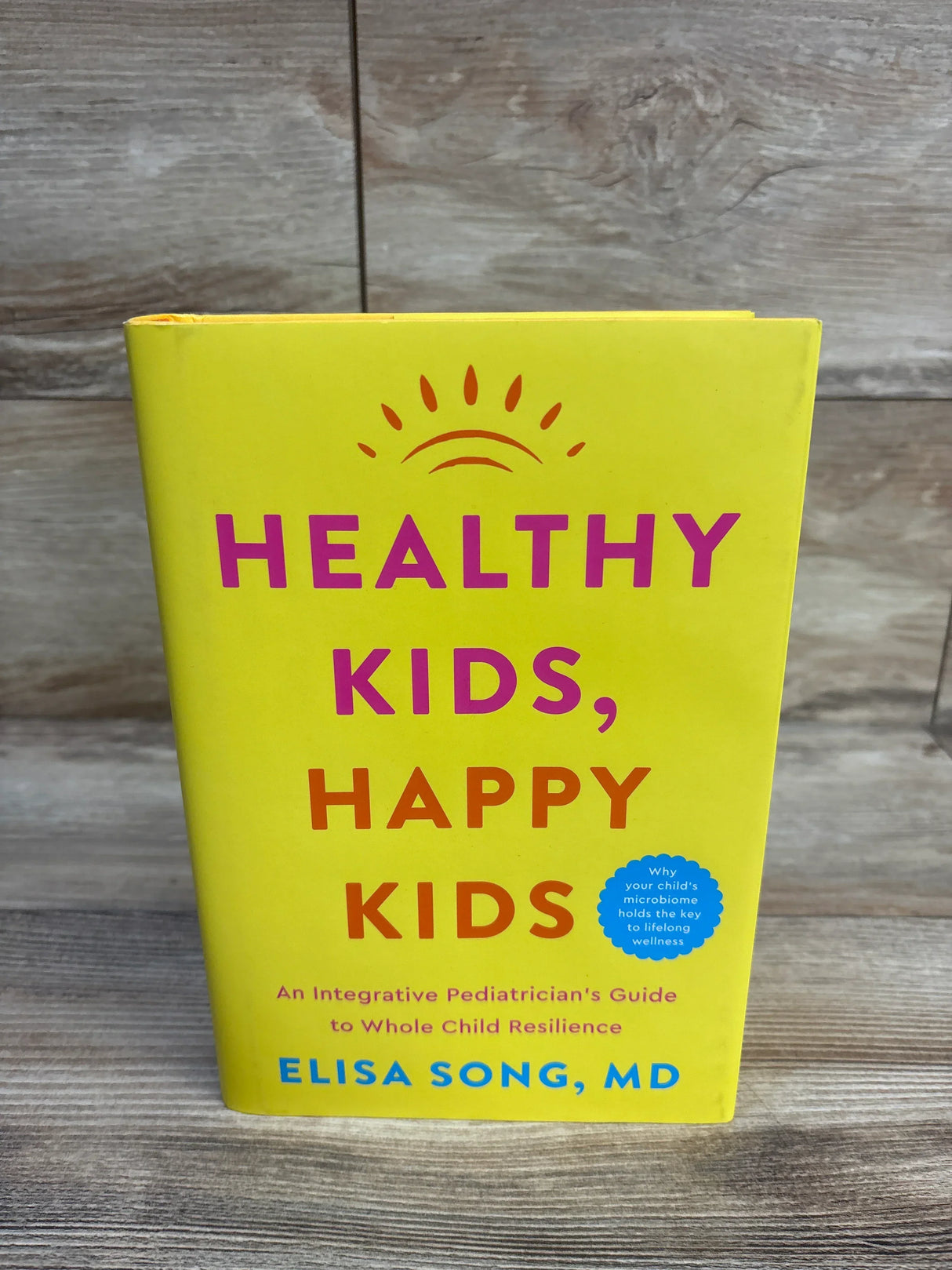 Front view of Healthy Kids, Happy Kids hardcover showing bright yellow cover with pink-orange title on wood background