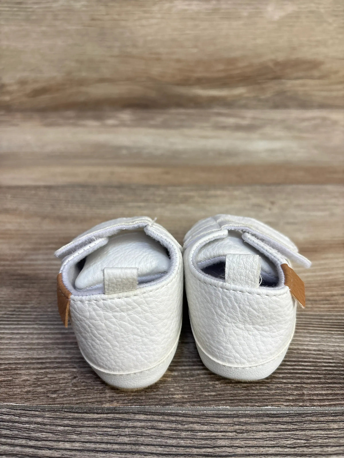 Rear view of Myggpp First Walker baby sneakers with white pebbled leather, Velcro straps, tan heel tabs on wood background