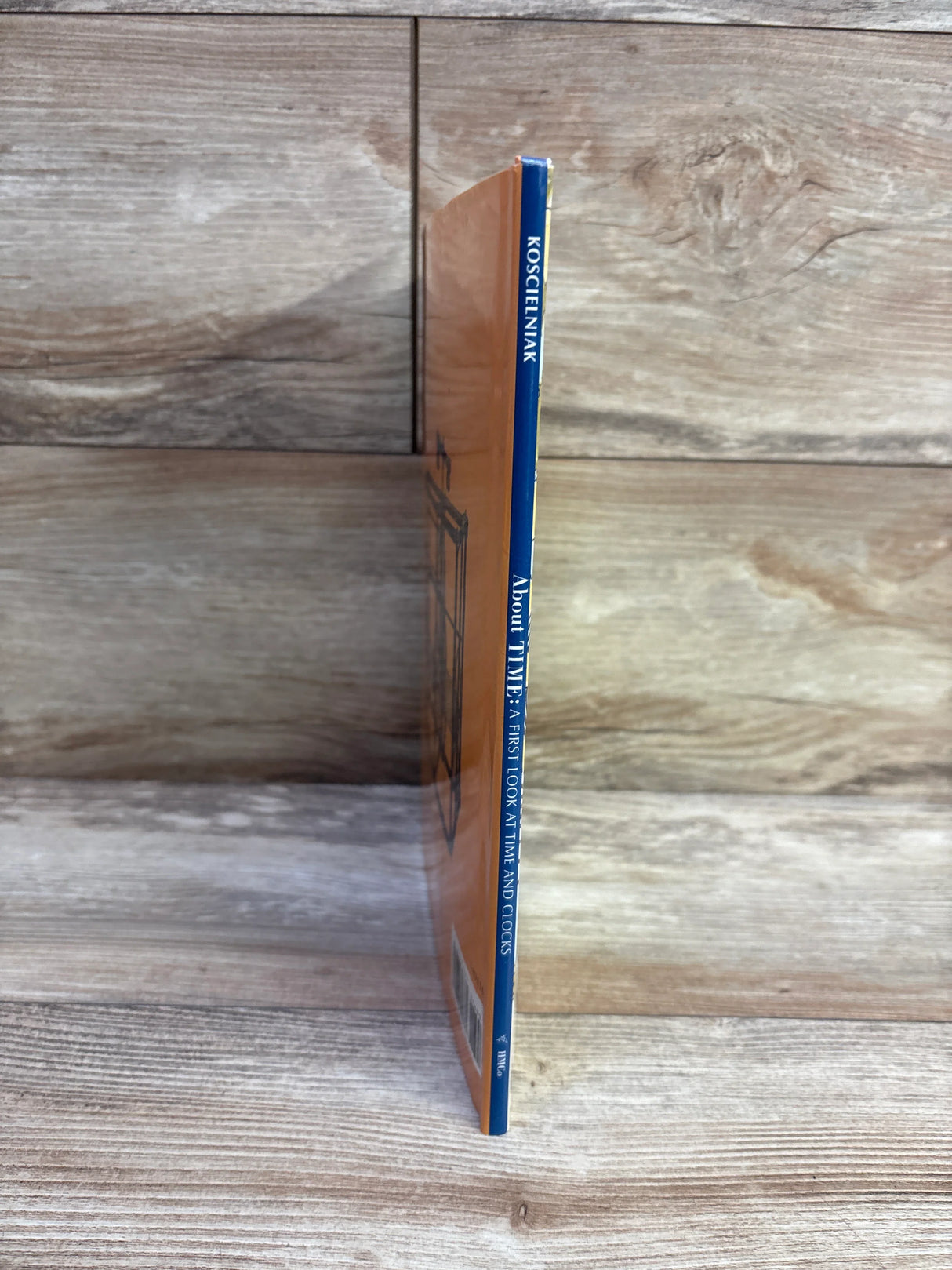 Side view of About Time hardcover book showing blue spine and orange cover against a wood background