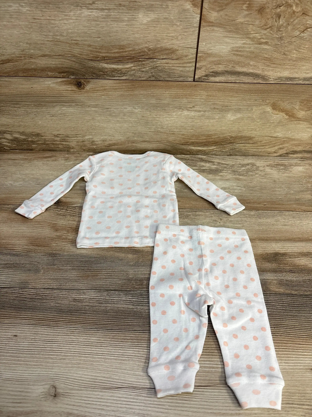 Flat lay of white baby two-piece pajamas with pale pink polka dots, cuffed long sleeves and ankle cuffs on wood floor
