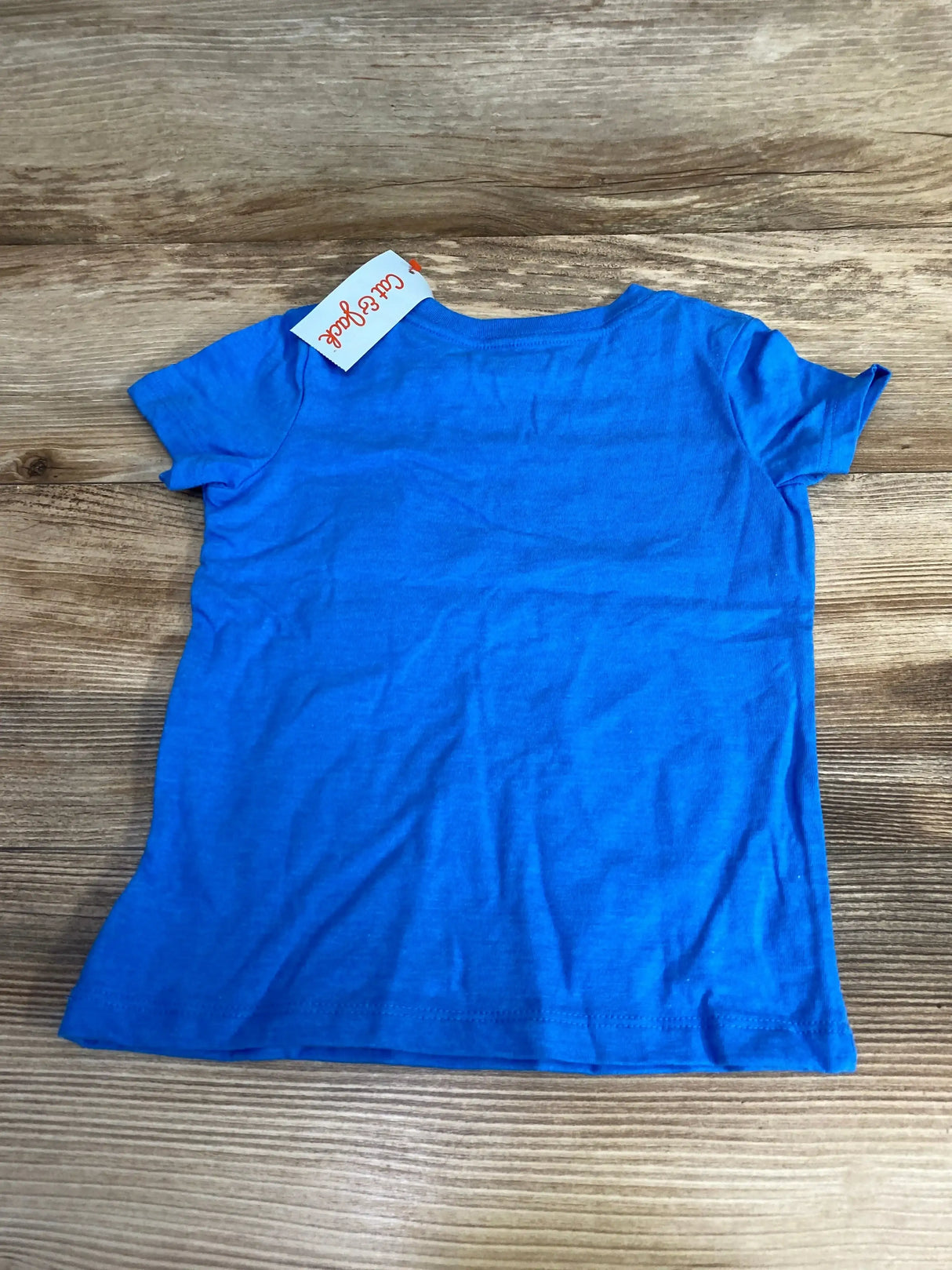 Back view of NWT Cat & Jack Dino Shirt size 2T showcasing solid blue short sleeve tee with brand hangtag on wood plank floor
