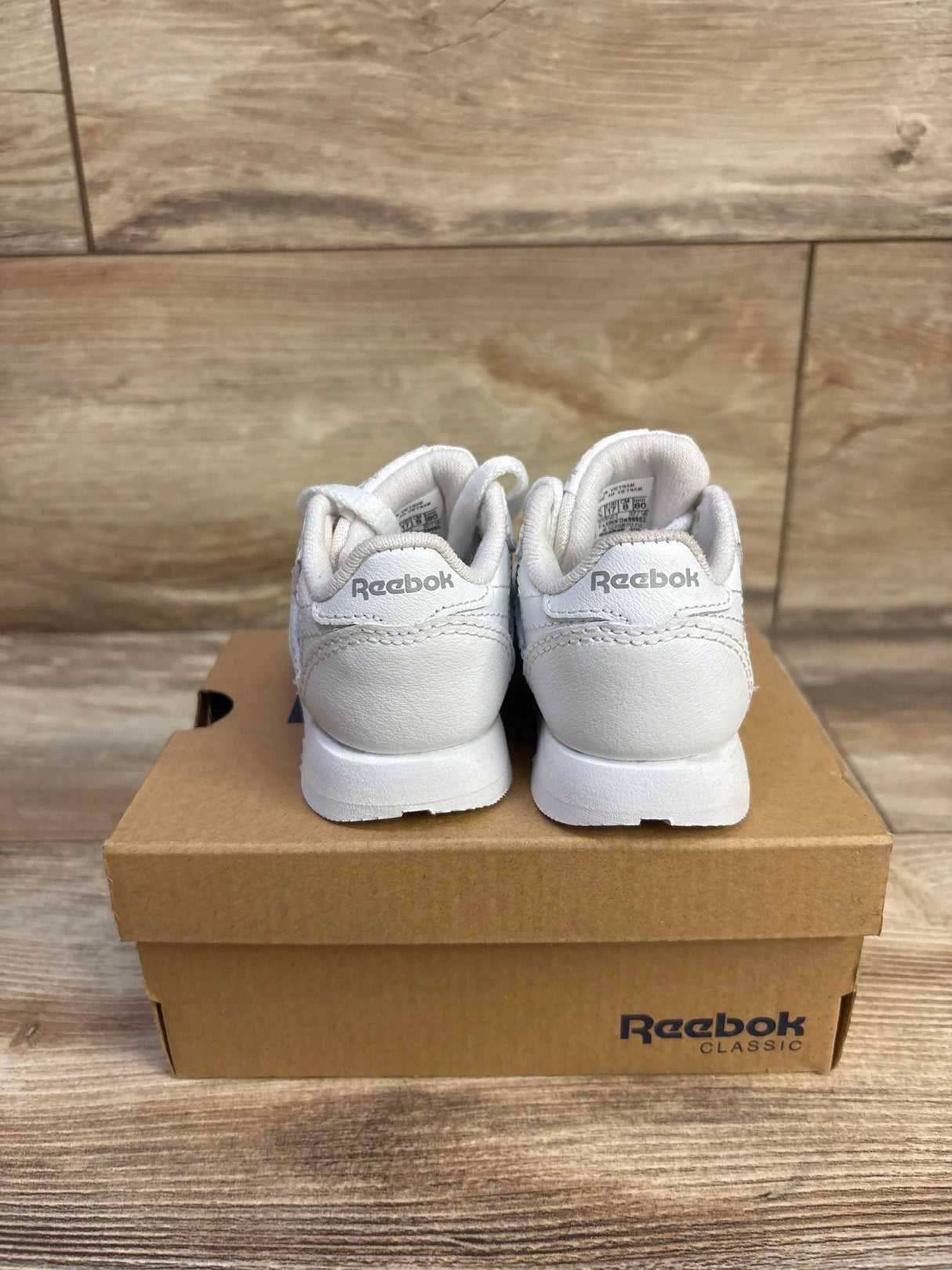 Rear view of white Reebok Classic Leather infant sneakers showing heel Reebok logo on a shoebox with wood background