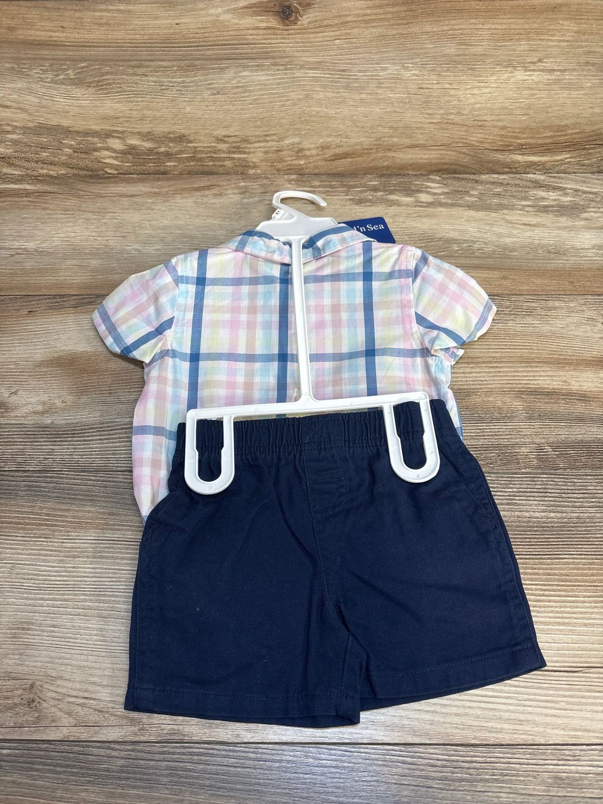 Front view of Land n Sea short-sleeve plaid bodysuit and navy shorts on hanger showing elastic waistband, wood background
