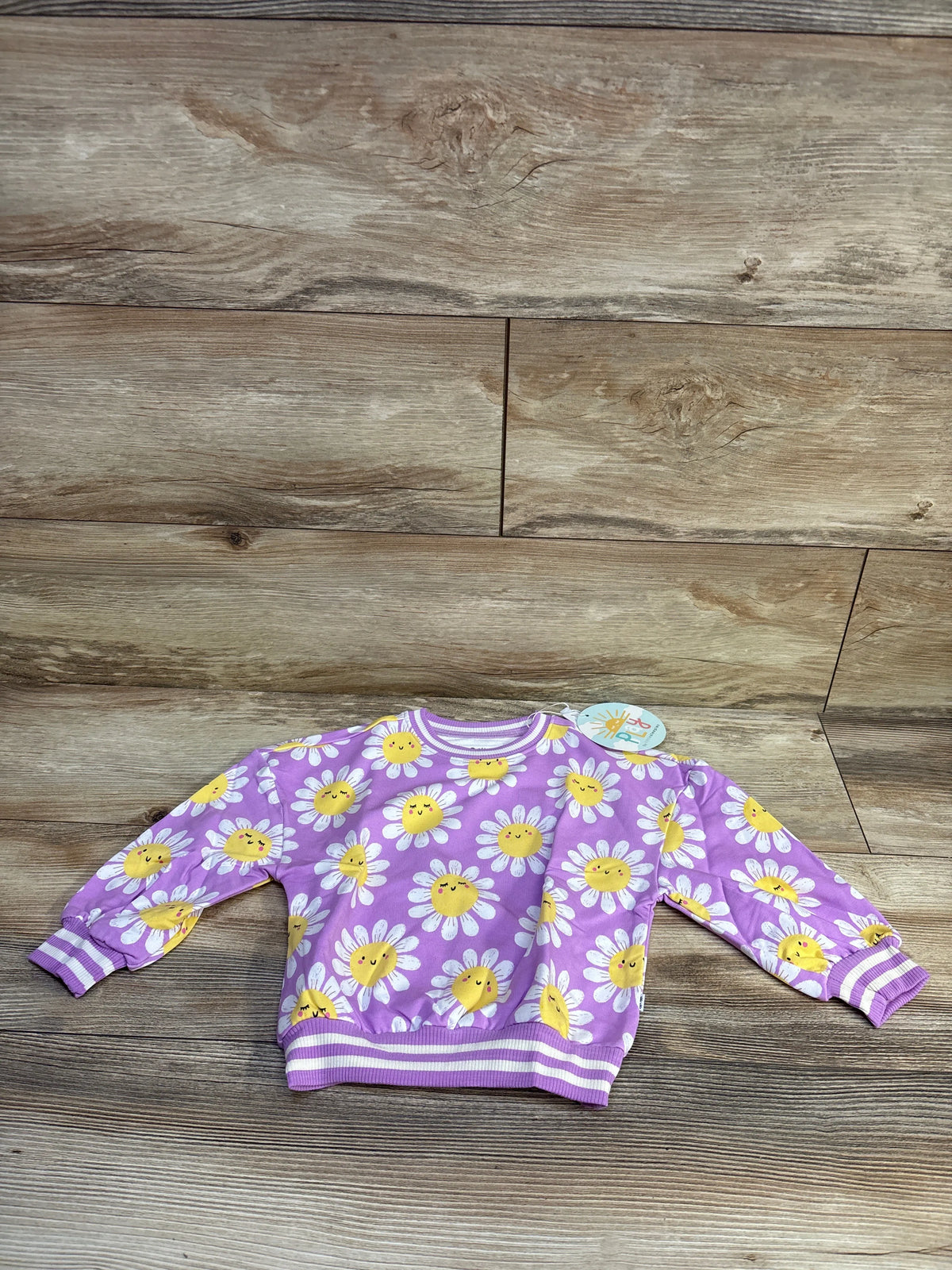 Front view of Little Sleepies purple bamboo pullover with smiling yellow daisy print and striped cuffs on wood background