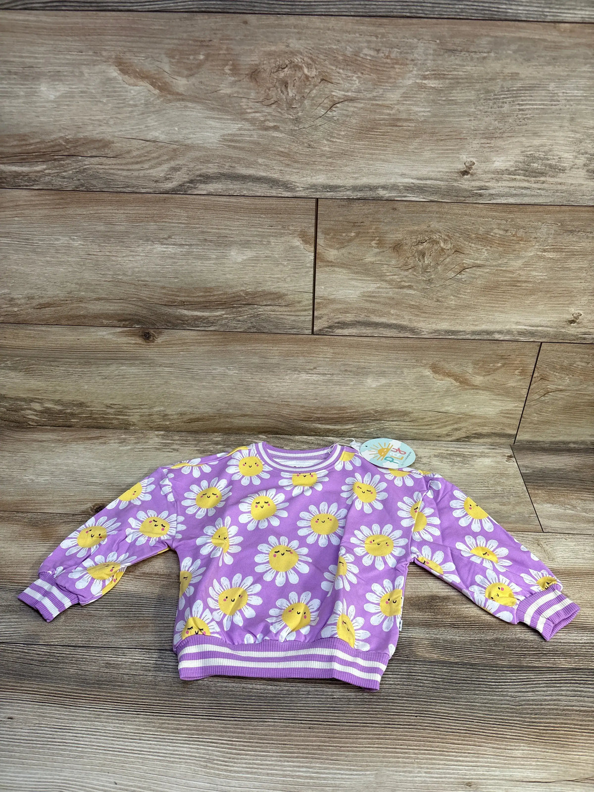 Front view of Little Sleepies purple bamboo pullover with smiling yellow daisy print and striped cuffs on wood background