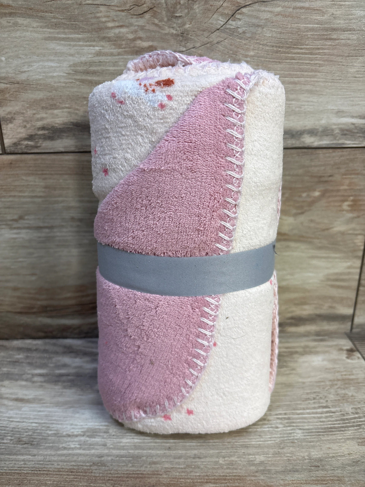 Rolled view of Cloud Island unicorn baby blanket in pink and cream with scalloped stitched edge and gray ribbon on wood background