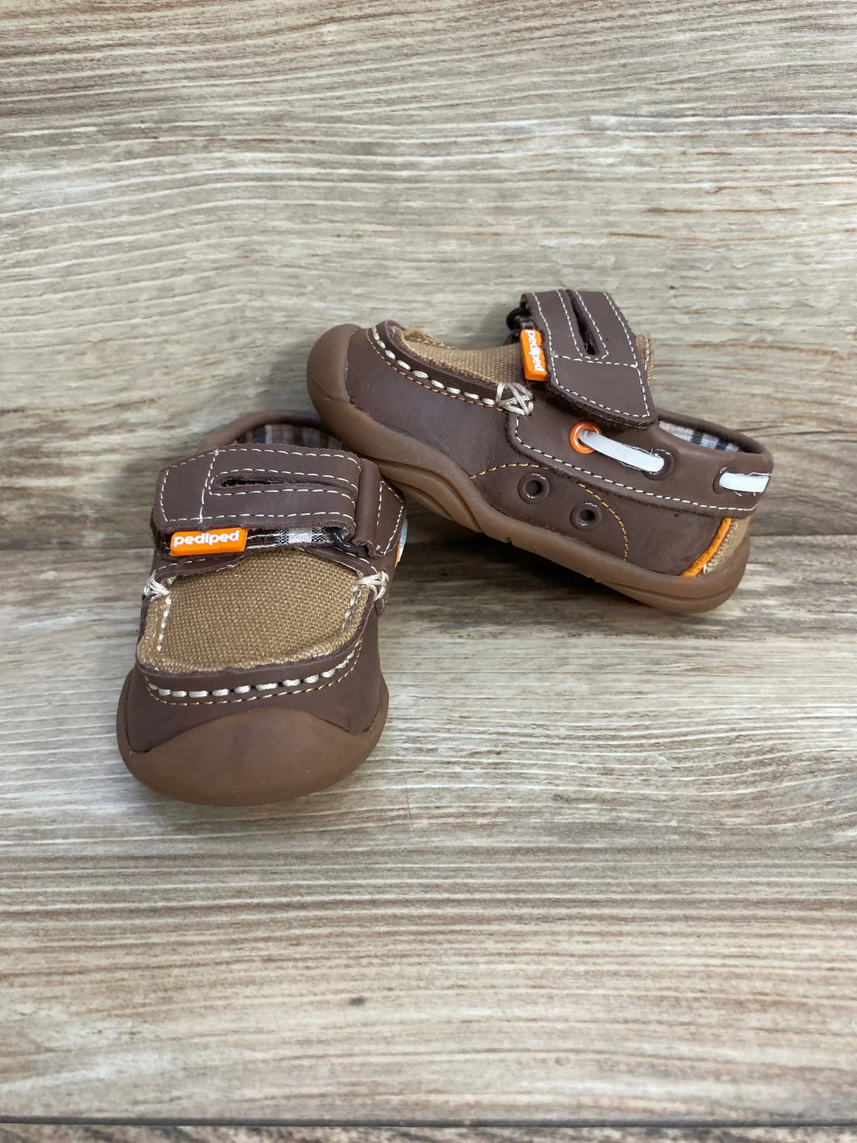 Angled front and side view of brown Pediped toddler loafers with Velcro strap, contrast stitching and rubber toe on wood