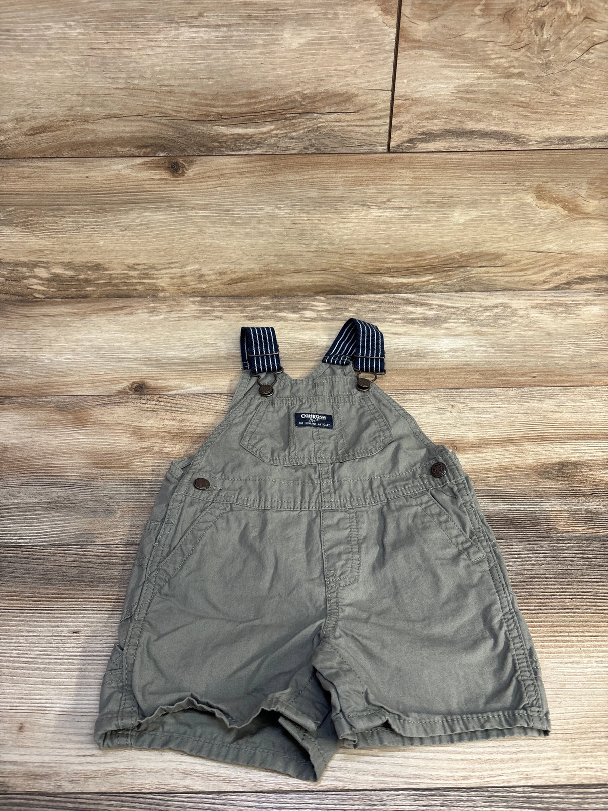 Flat lay of green OshKosh shortalls 18 months with striped adjustable straps, front bib pocket and side snap buttons on wood