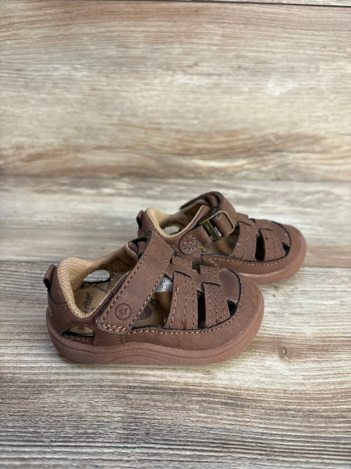 Three-quarter view of brown Stride Rite Amos 3.0 fisherman sandals with adjustable straps on a wood-grain surface