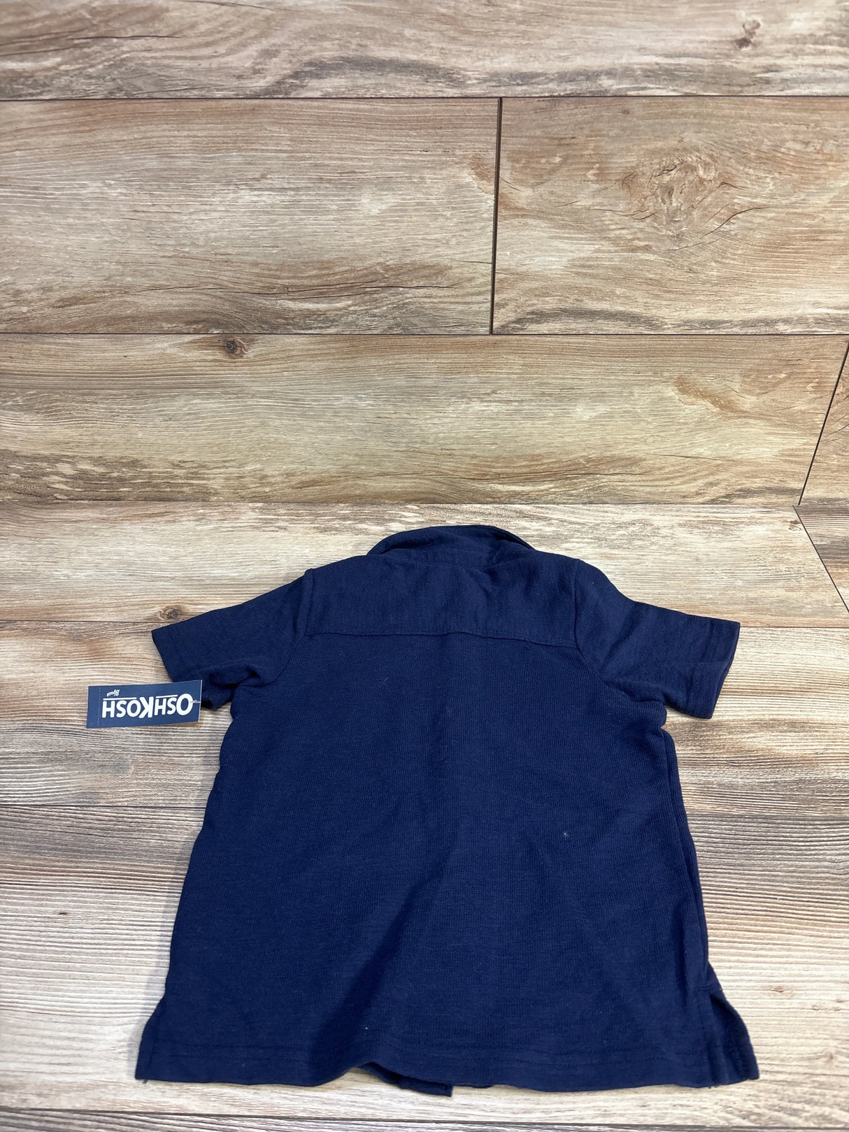 Back view of Oshkosh navy short-sleeve button-up toddler shirt size 4T showing back yoke and attached brand tag on wood plank background