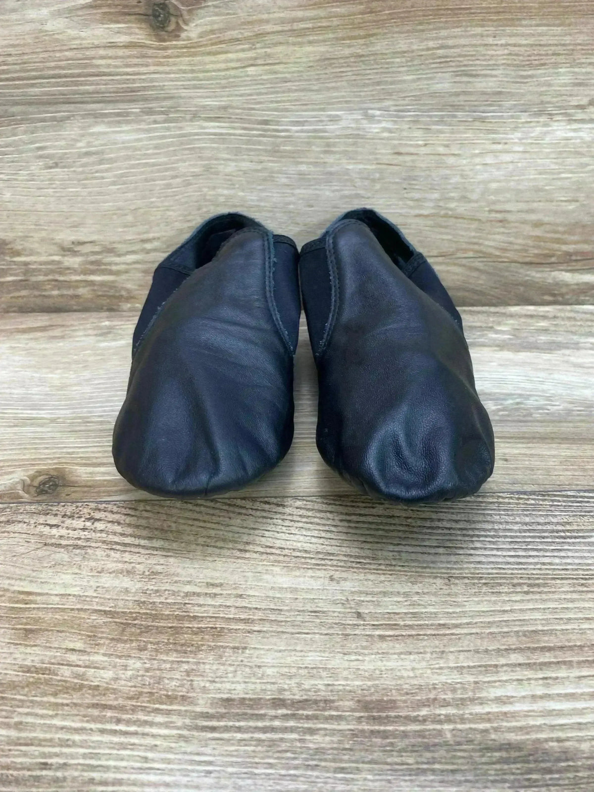 Front view of Capezio E-Series black leather jazz slip-on shoes with elastic side panels on light wood background