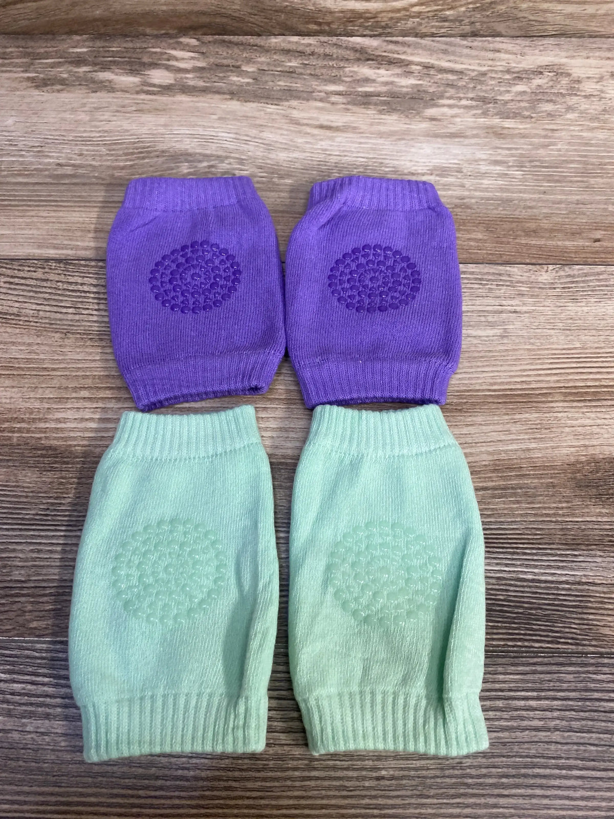 Top-down view of two pairs of baby crawling knee pads in purple and mint showcasing silicone grip dots and elastic cuffs on a wood surface
