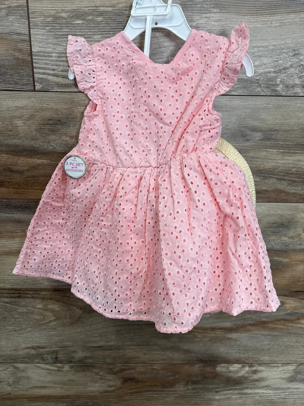 Pink eyelet baby dress with ruffled sleeves, displayed on a hanger, wood background