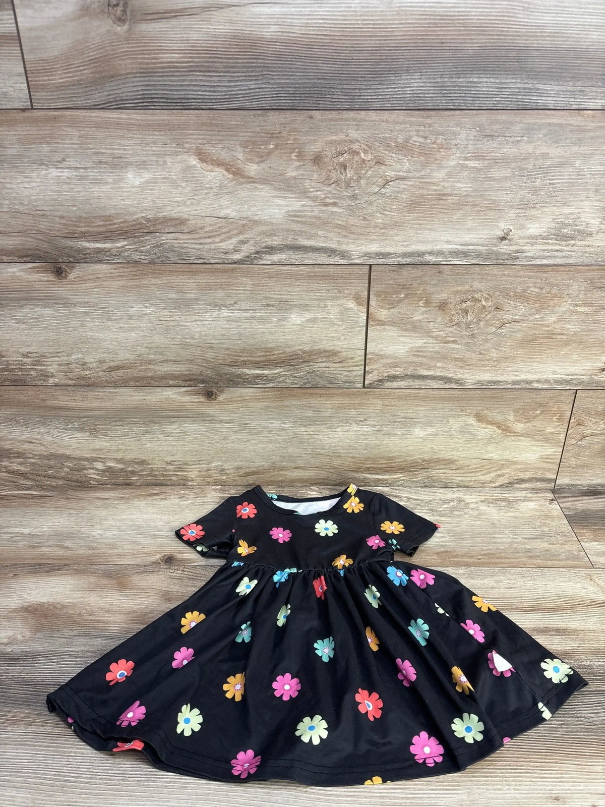 Flat lay of black toddler short-sleeve dress with colorful daisy print and gathered waist, size 12-18 months, on light wood floor