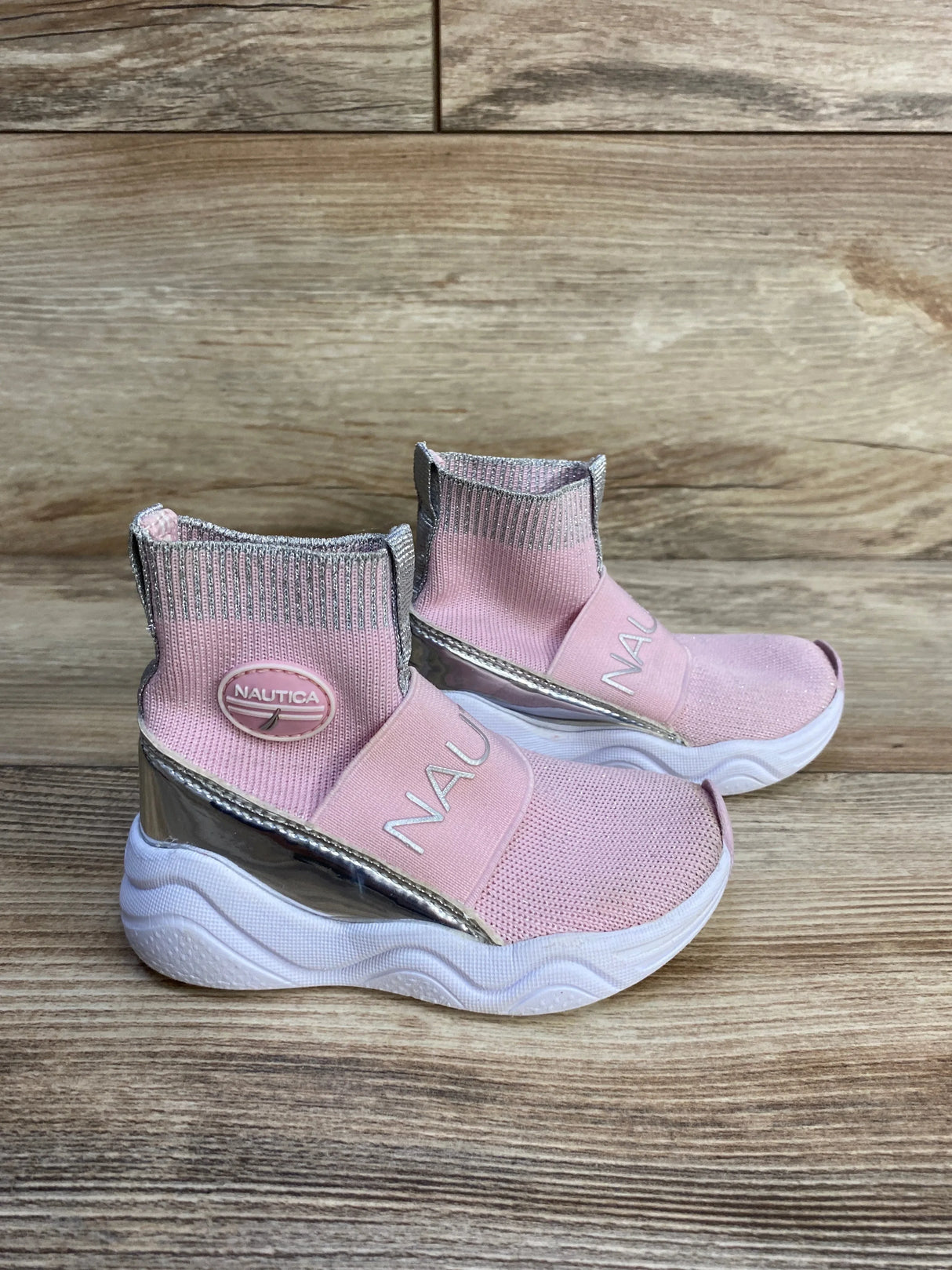 Side view of pink Nautica slip-on sneakers with knit ankle, NAUTICA strap, silver heel trim and white wavy sole on wood