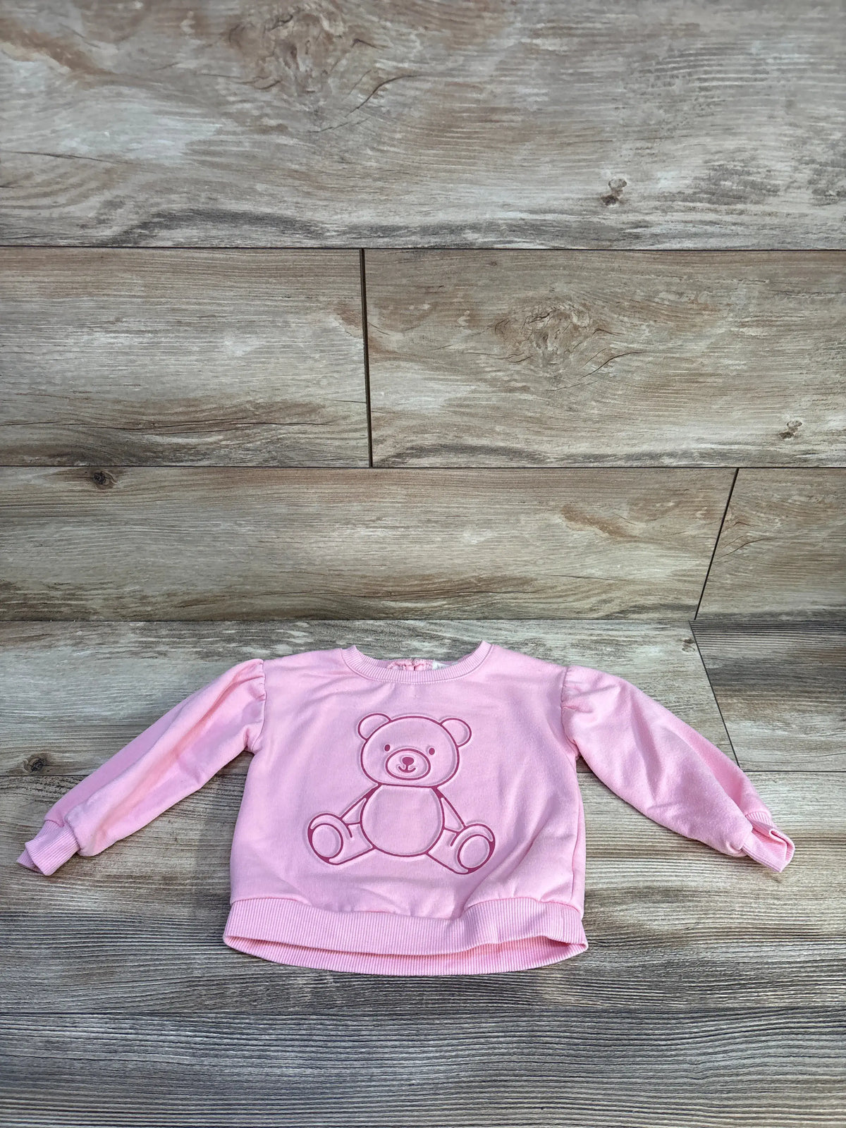 Front view of Btween bear sweatshirt in pink showcasing a teddy bear design on a wood floor