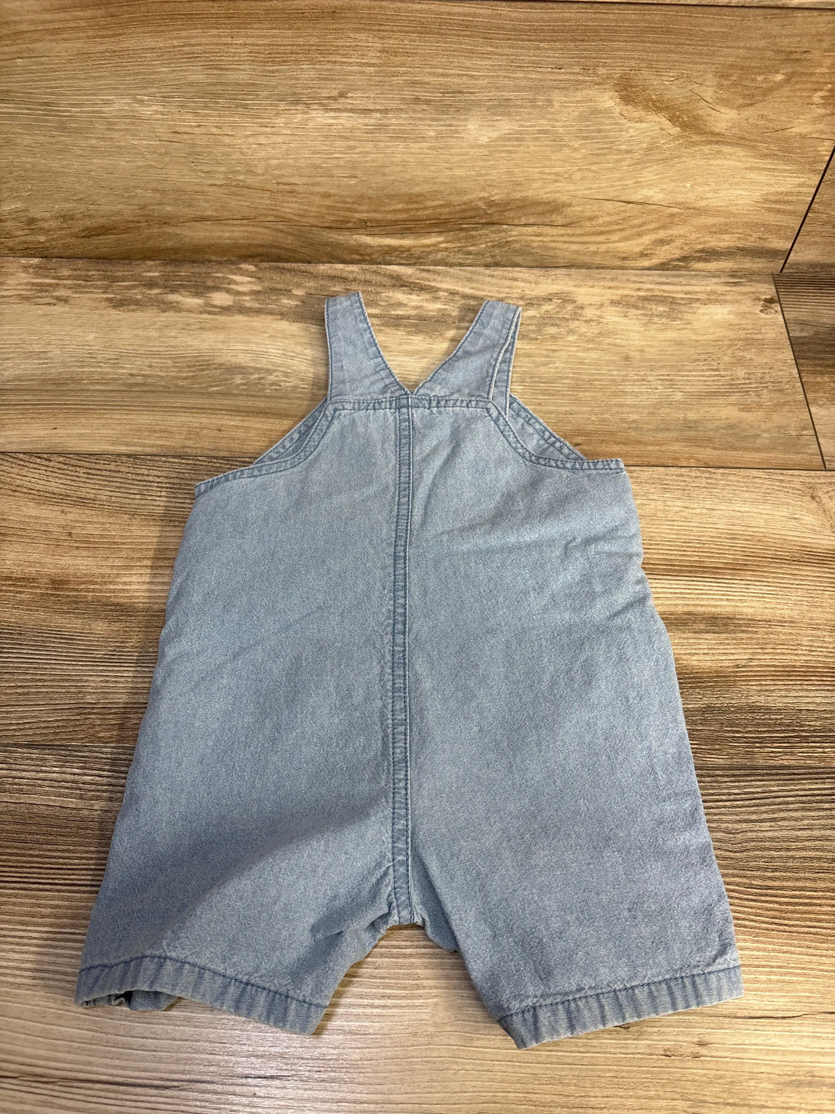 Front view of Carter’s blue chambray shortalls in size 9 months showcasing adjustable straps on a wood-grain background