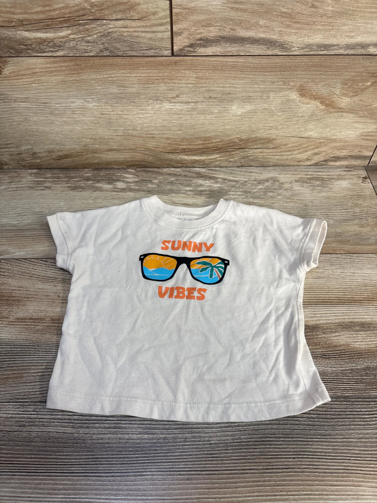 Front view of ivory Okie Dokie Sunny Vibes shirt in size 12 months with sunglasses graphic on wood grain