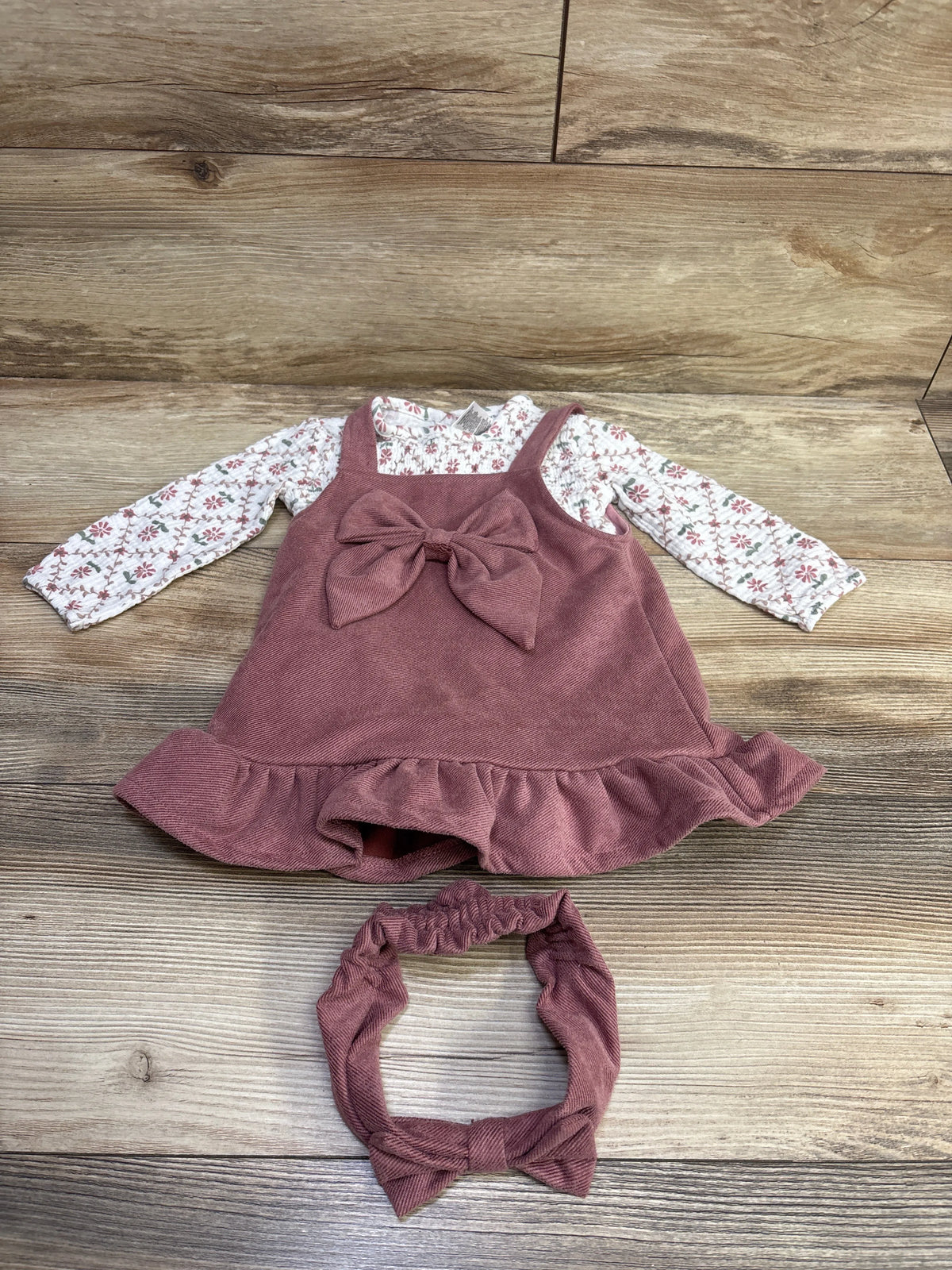 Top view of Daisy Fuentes 3-piece outfit with white floral bodysuit, mauve skirtall with bow, and matching headband on wood