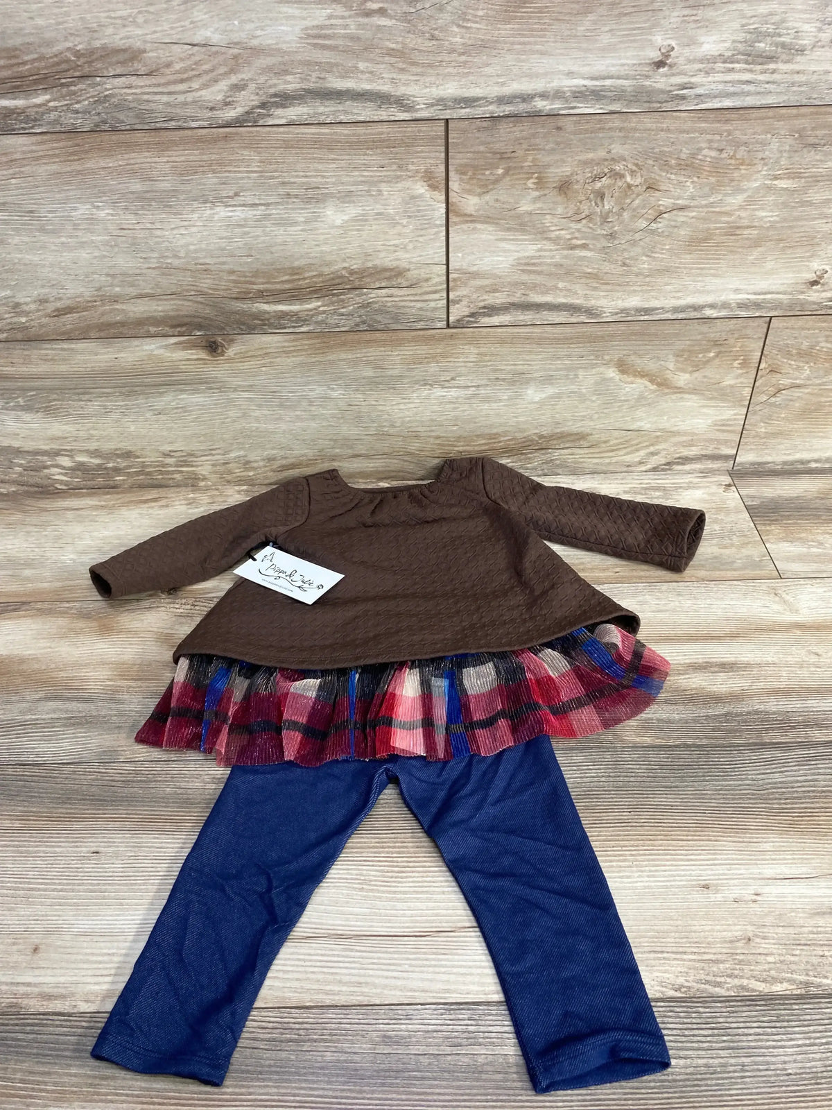 Front view of 3-piece toddler sweatshirt and bottoms set with brown textured top, plaid ruffle skirt, and blue leggings on wood floor