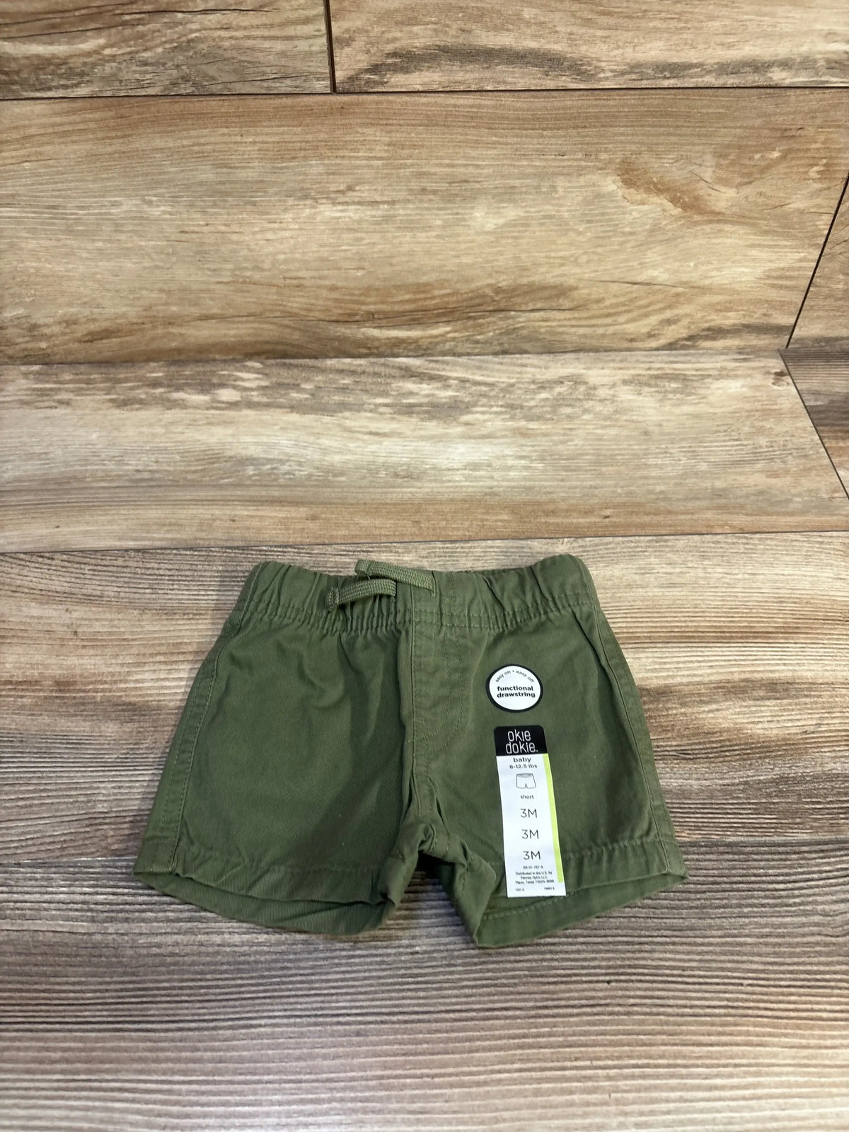 Top-down front view of green Okie Dokie baby drawstring shorts showing elastic drawstring waistband and size tag on wood plank background