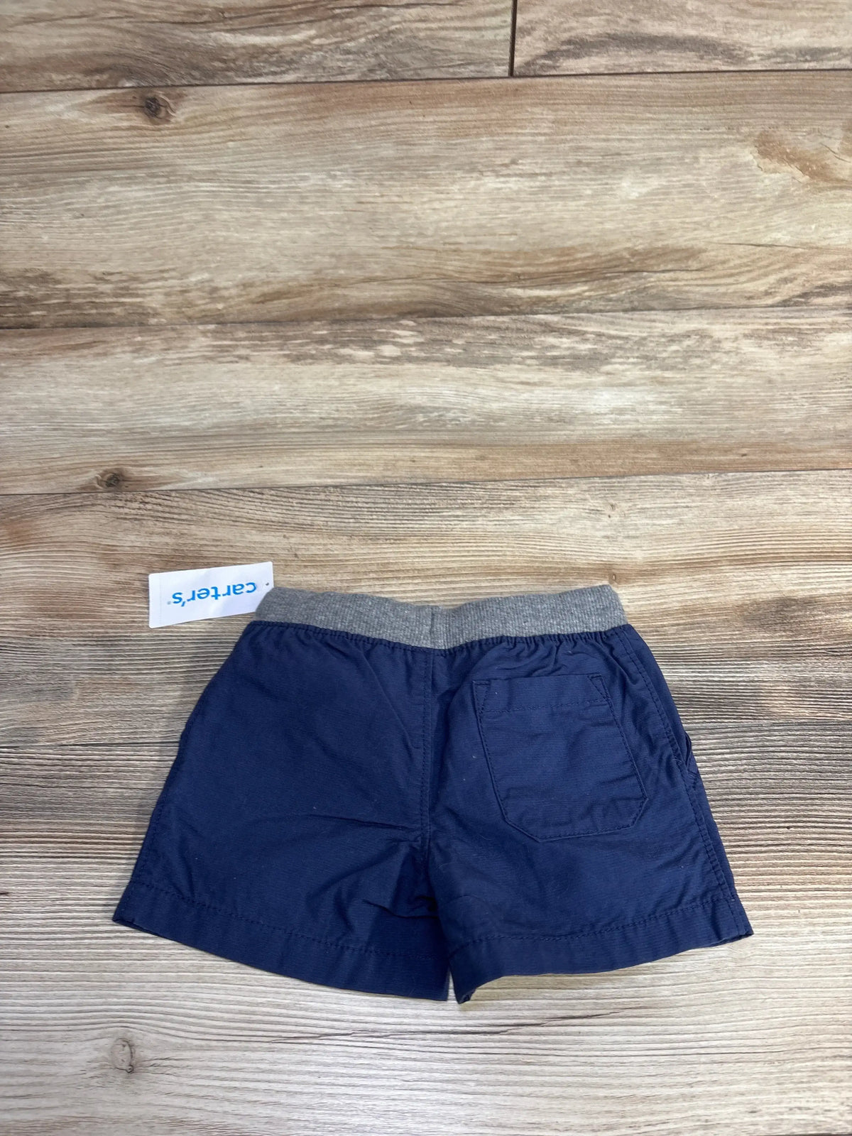Back view of Carter’s navy shorts 6m showcasing gray elastic drawstring waistband and back patch pocket on wood background