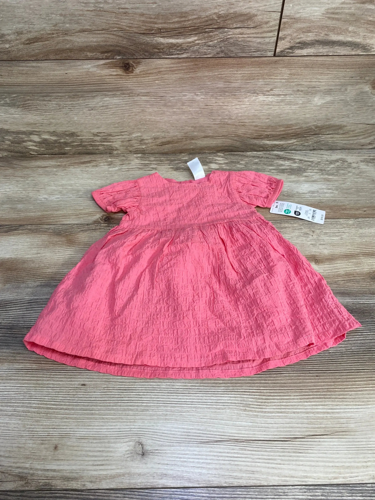 Flat lay of pink baby dress showing crinkled texture, short puff sleeves, and attached size tag on wood-grain background