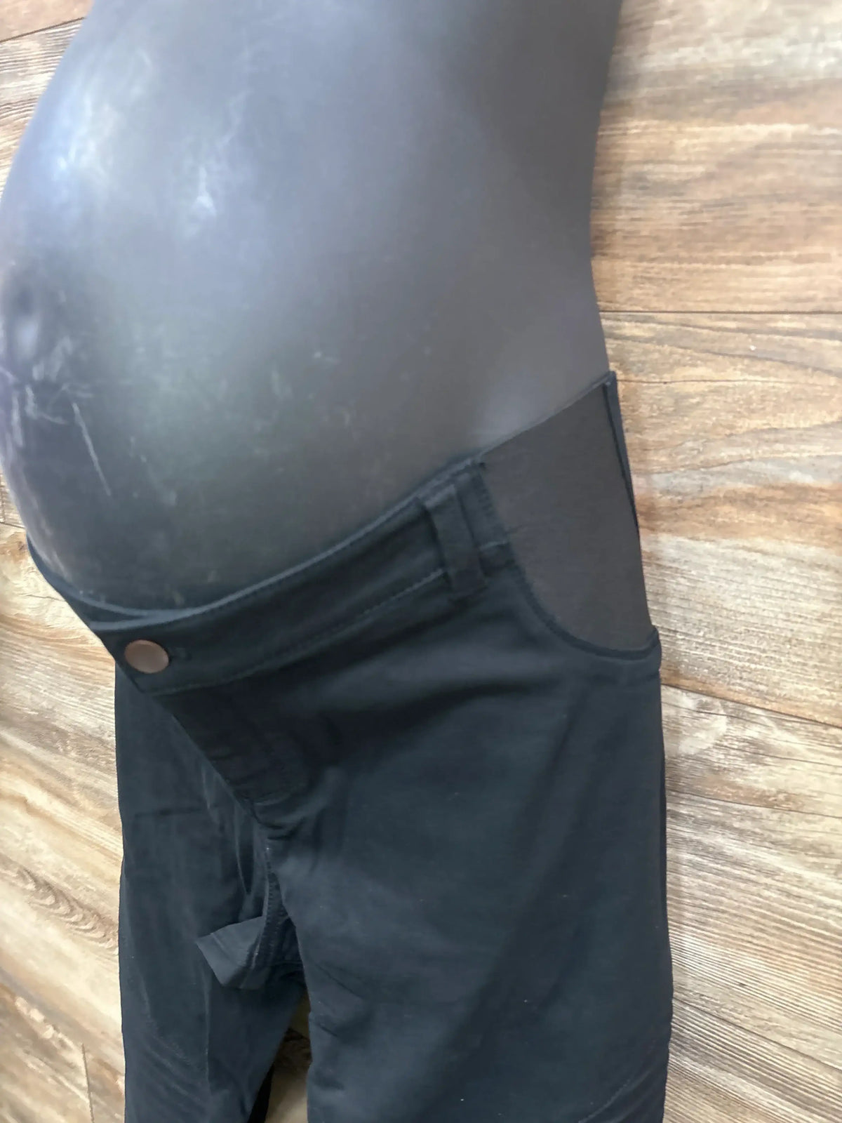 Side view of Isabel maternity utility jeggings in black showing stretch belly panel, front button and pocket on wood floor background