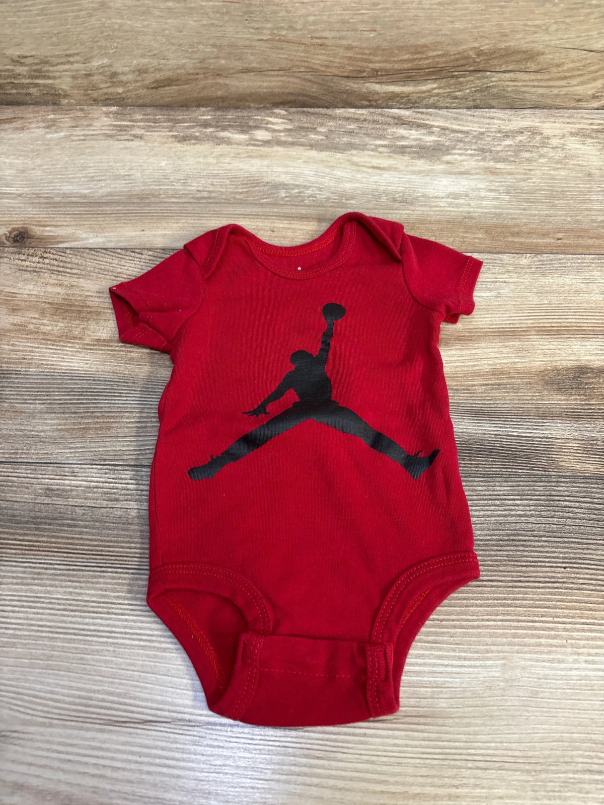 Flat-lay of red baby bodysuit showcasing black Jumpman logo, short sleeves, and three-snap crotch on wood background