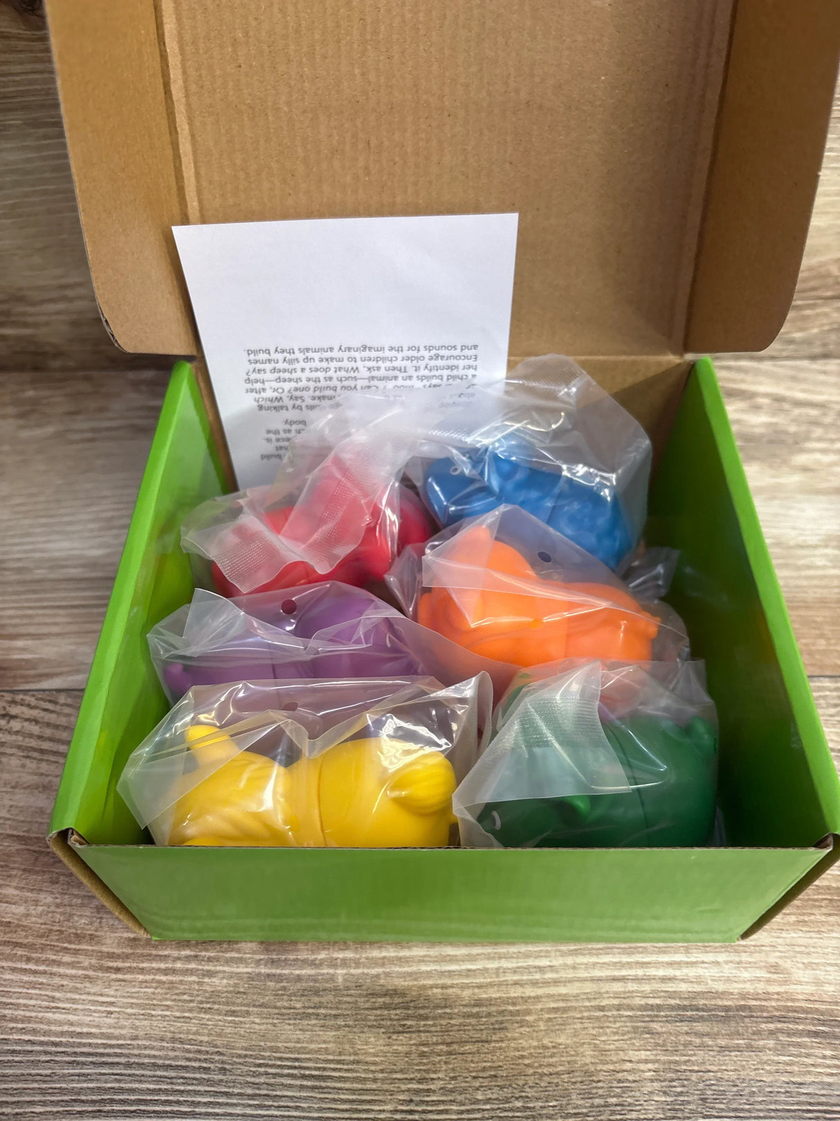 Top view of NWT Lakeshore Mix and Match Magnetic Farm Animals in open green box with colorful bagged pieces and instruction sheet on wood plank floor