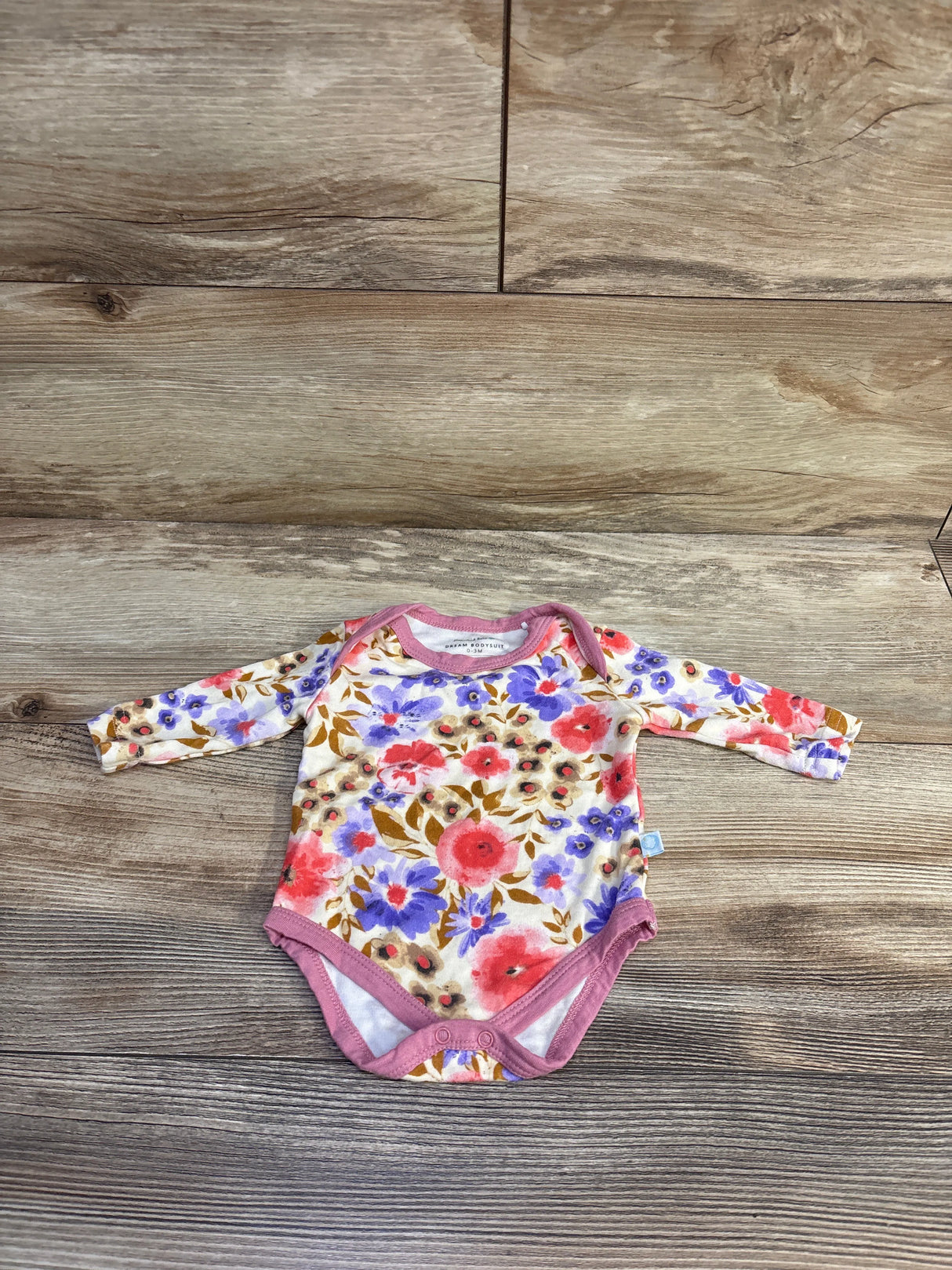 Flat lay of long-sleeve Dreamland Baby bamboo floral bodysuit showing cream background, pink trim, and pink and purple flowers on wood backdrop
