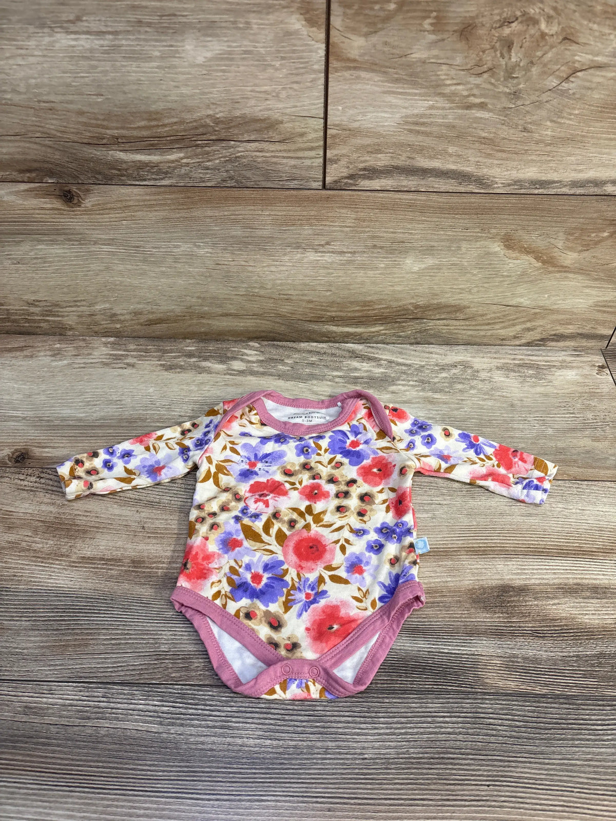 Flat lay of long-sleeve Dreamland Baby bamboo floral bodysuit showing cream background, pink trim, and pink and purple flowers on wood backdrop