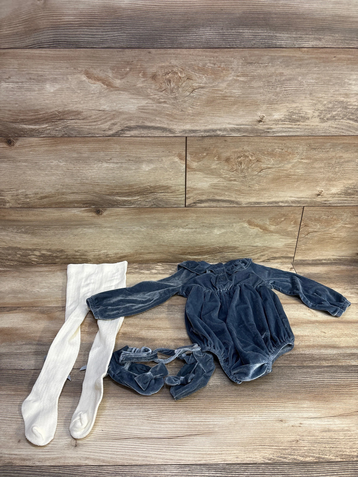 Flat lay of blue 3-piece velvet romper set with long-sleeve romper, matching bow booties and white ribbed tights on wood