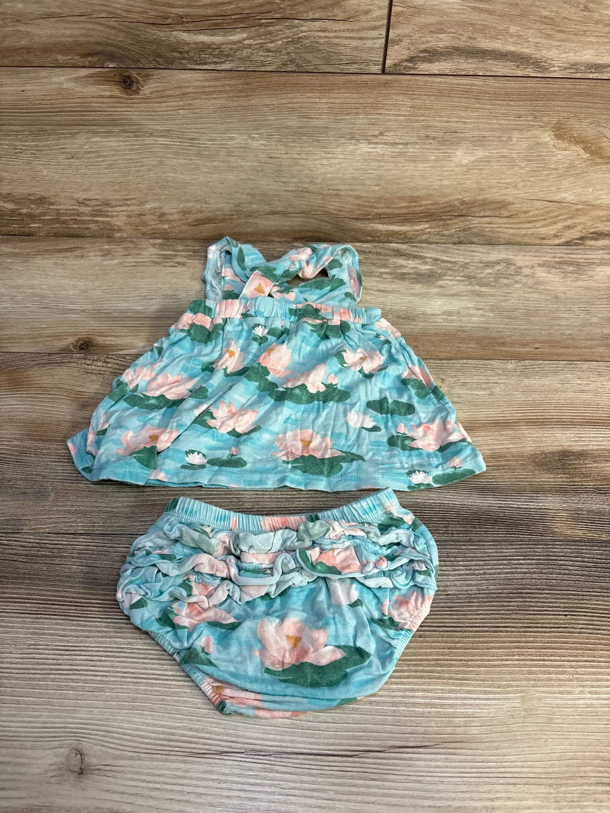 Flat lay of Angel Dear baby pinafore dress and matching bloomers showing pink lily pad floral print on blue fabric on wood background