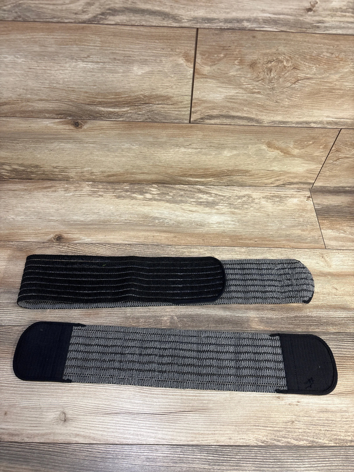 Top view of Belly Bandit 2-in-1 Hip Bandit in black and gray, size XS-M, showing elastic ribbed support panels and Velcro closures on wood floor
