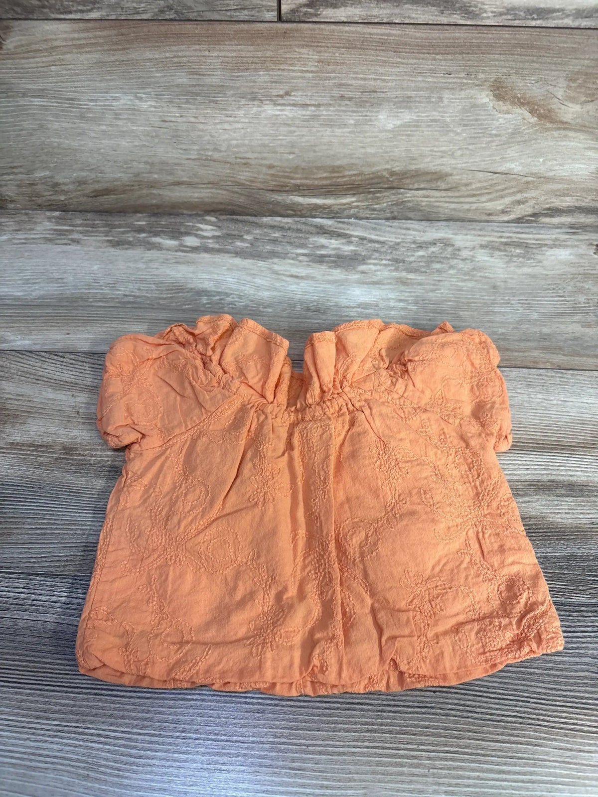 Front view of orange toddler cold-shoulder top showcasing floral embroidery and ruffled neckline on light wood background