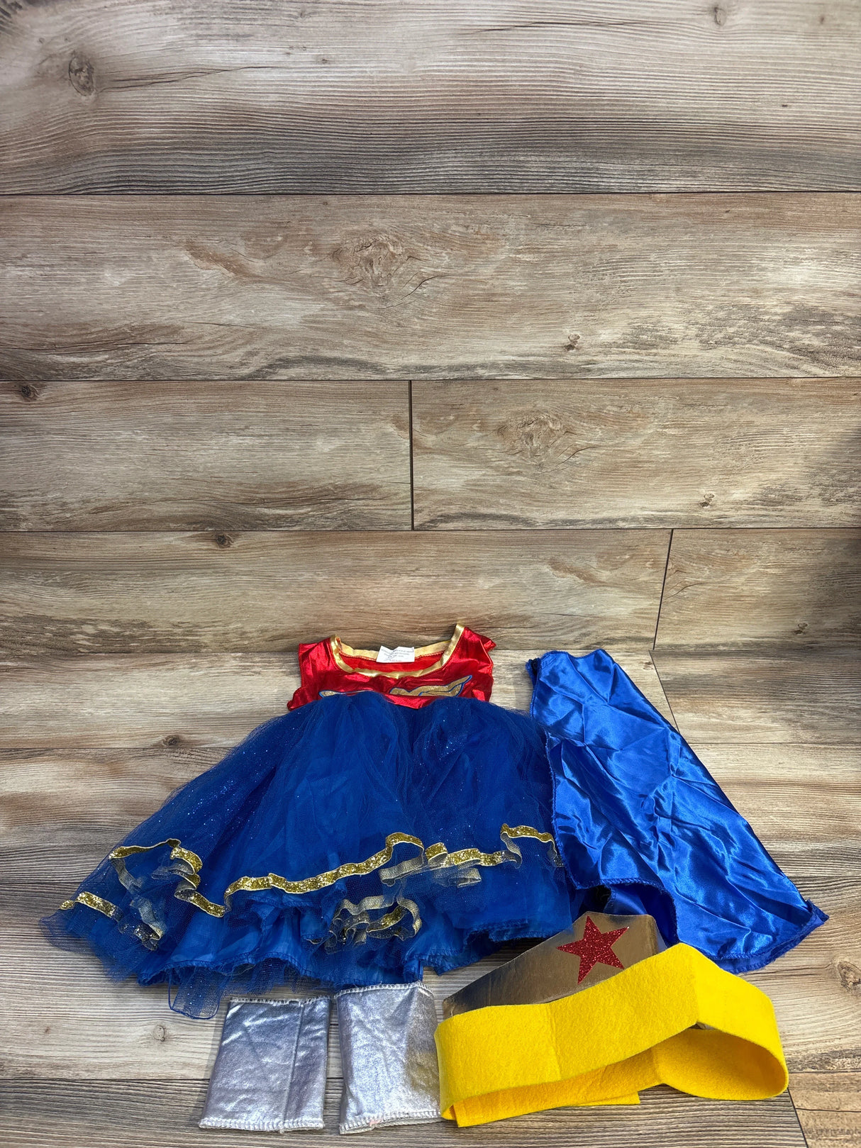 Flat lay of Wonder Woman toddler costume with red top, blue tulle skirt, cape, silver boot covers, and yellow headband on wood