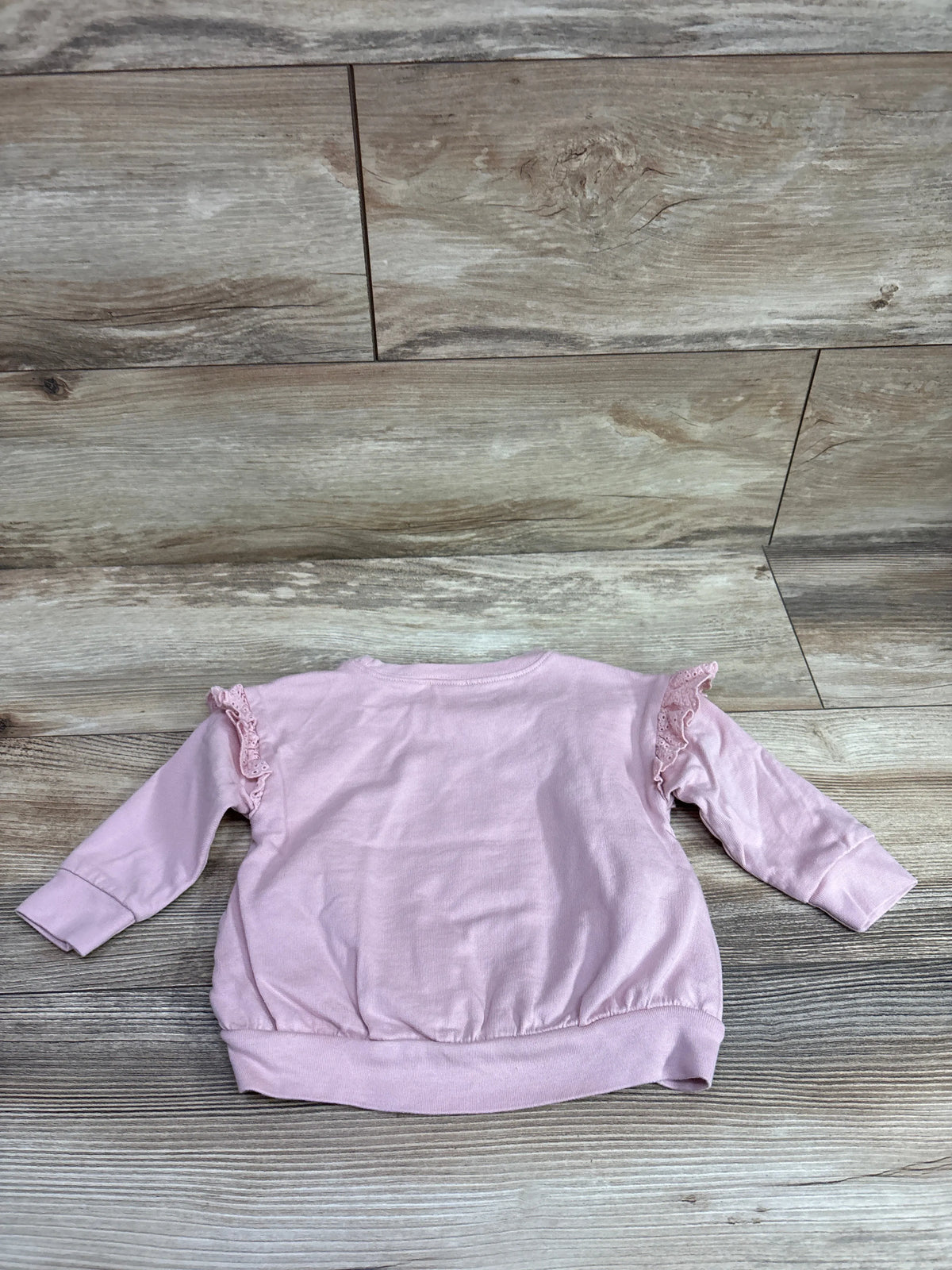 Front view of pink Just One You sweatshirt in size 12 months with lace ruffle shoulders and ribbed cuffs on a wood floor