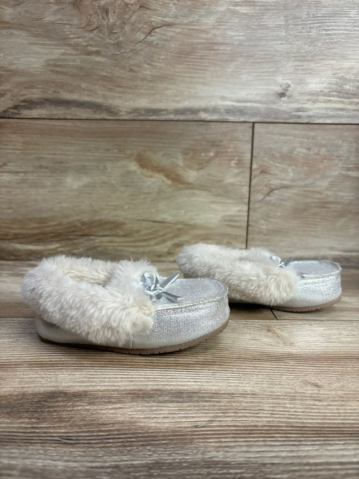 Pair of silver toddler moccasin slippers with soft cream faux fur lining and bow detail on wooden floor