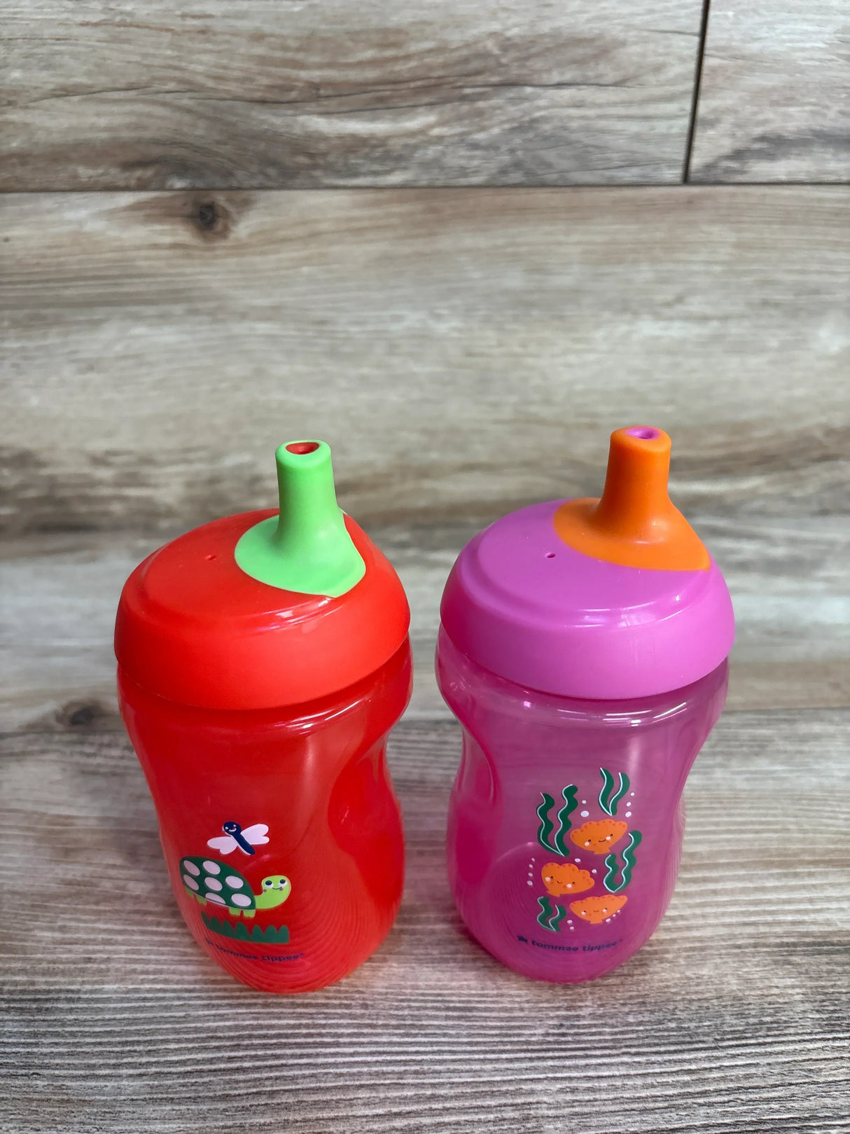 Two Tommee Tippee sippy cups, one red with turtle design, one pink with fish, on wood surface