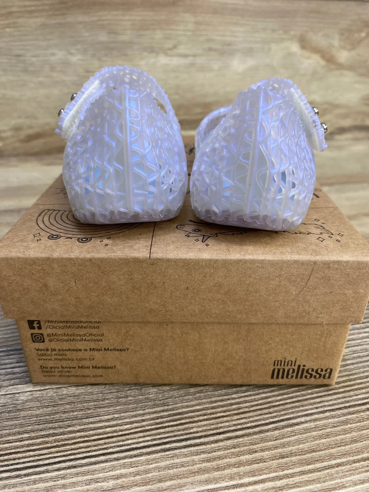 Rear view of Mini Melissa Campana Papel jelly flats showcasing pearly blue lattice-pattern heels on a brown Mini Melissa shoebox on wood surface