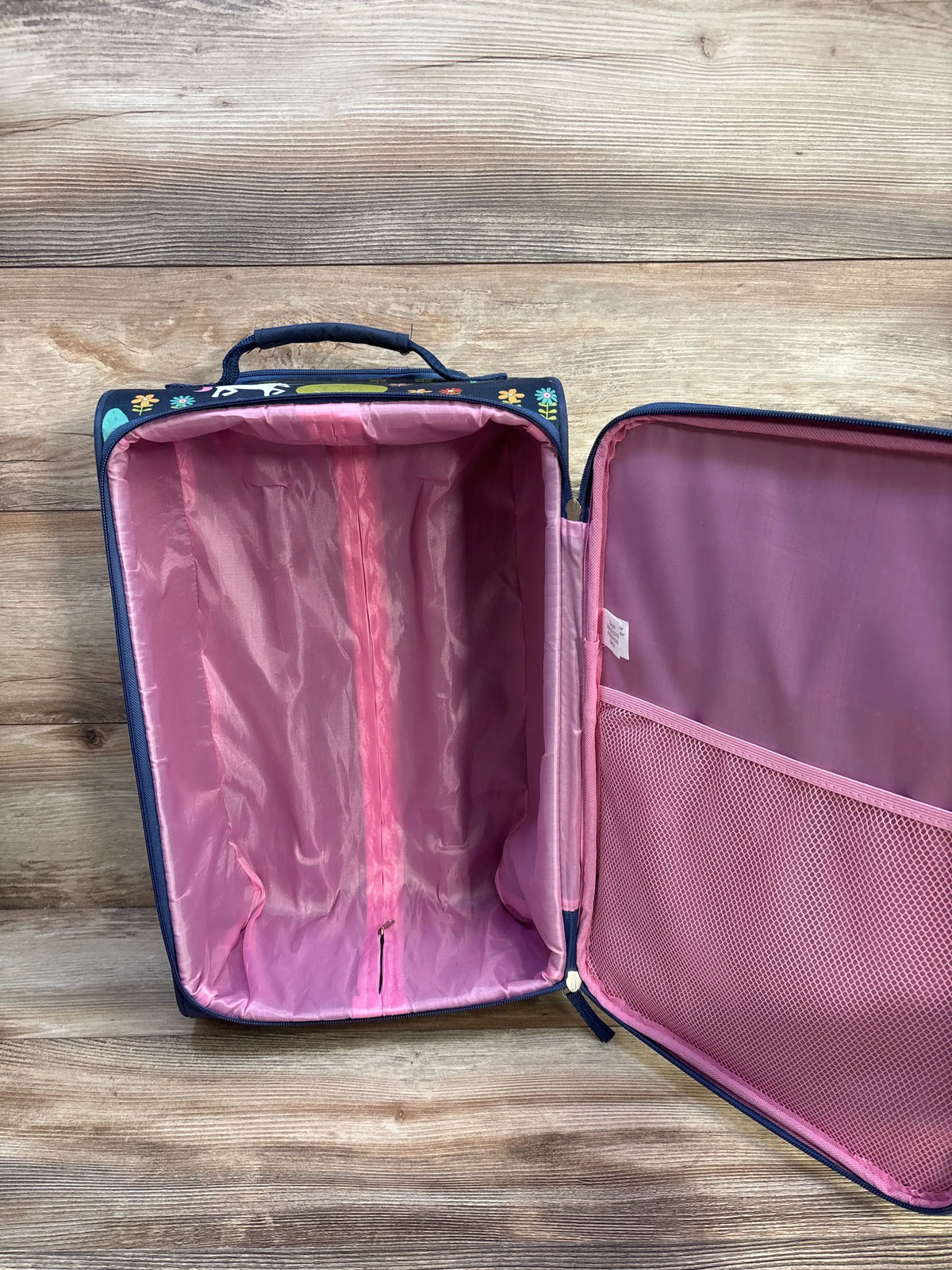 Open interior view of Crckt Kids’ Softside Carry On Suitcase showcasing pink lining and mesh pocket on wood floor
