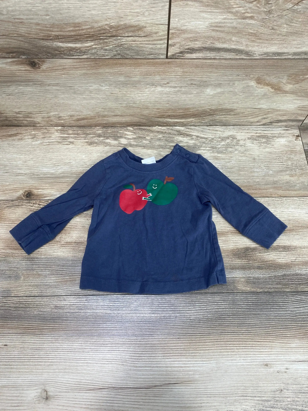 Front view of Hanna Andersson long-sleeve baby shirt with red and green apple print on wood background, size 6–12M