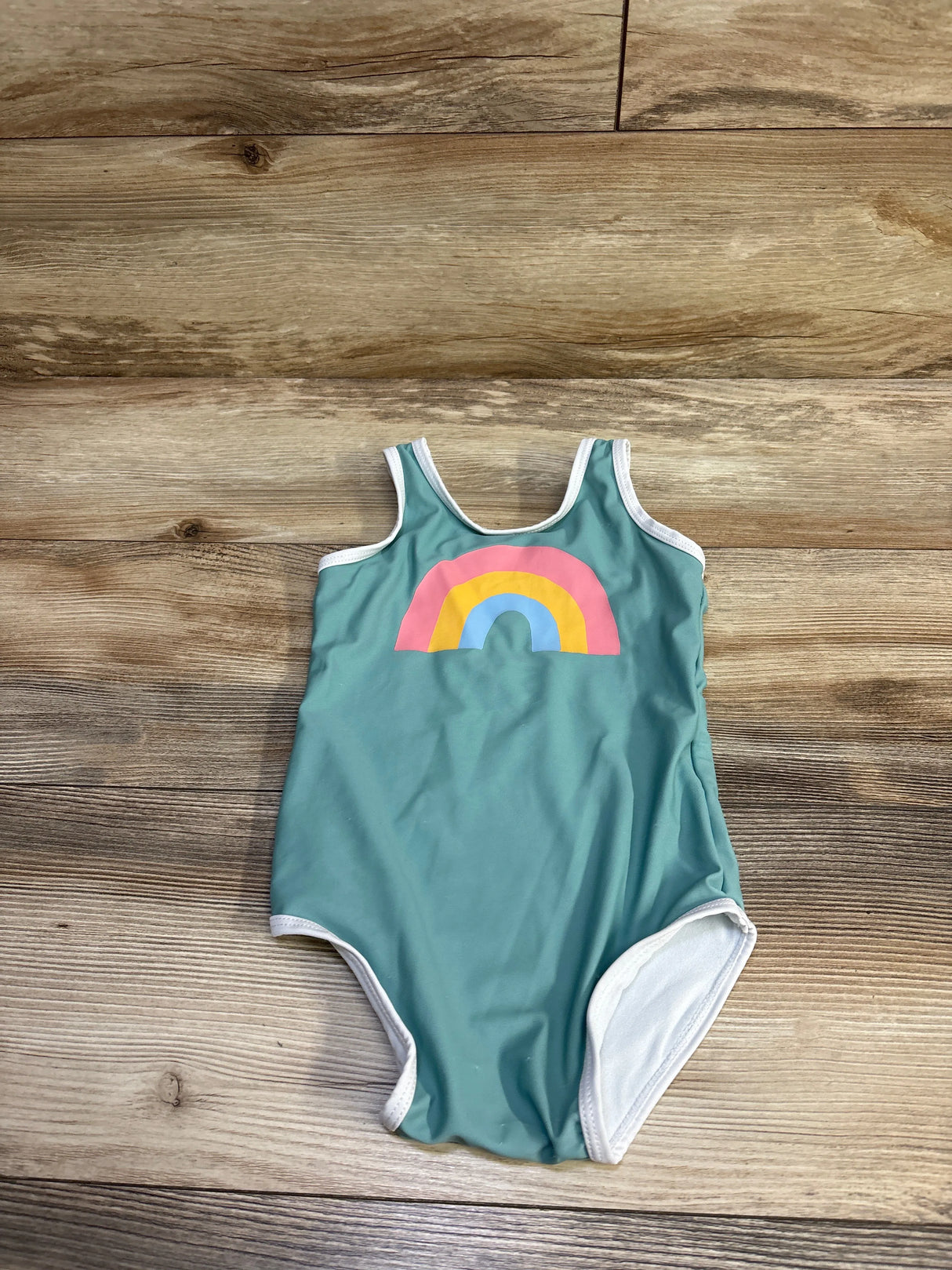 Flat lay of green one-piece baby swimsuit showing rainbow chest print and white trim on wood floor