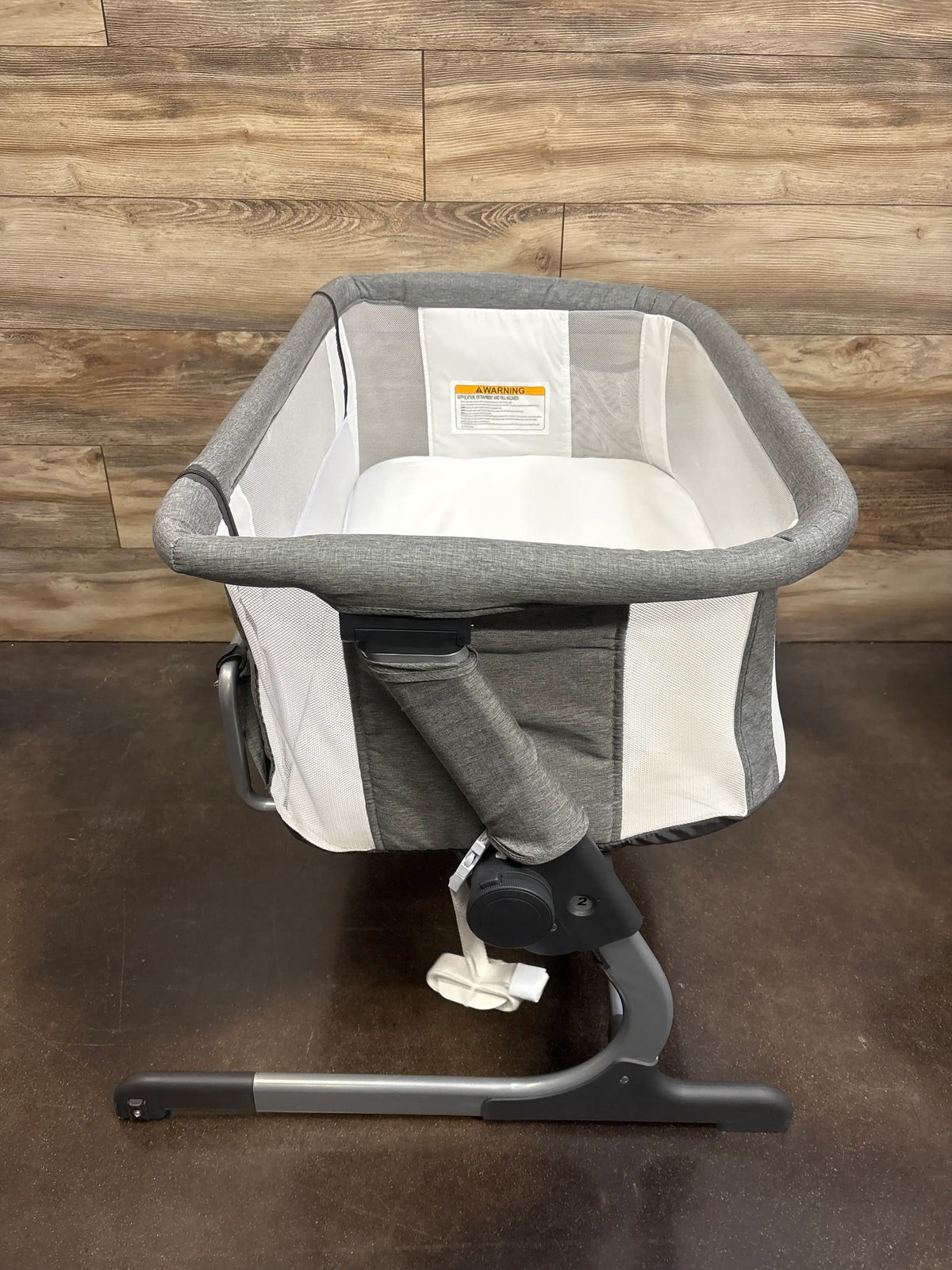 Front view of Baby Delight Beside Me Dreamer bedside bassinet in grey with mesh sides and adjustable base on a wood floor