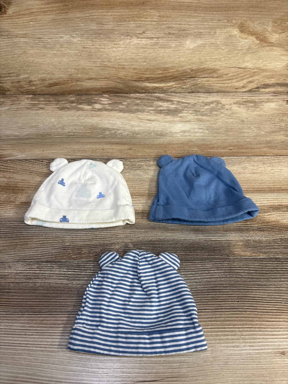 Top view of three Baby Gap organic cotton beanies in blue and white with bear ear appliques on a wood-grain background
