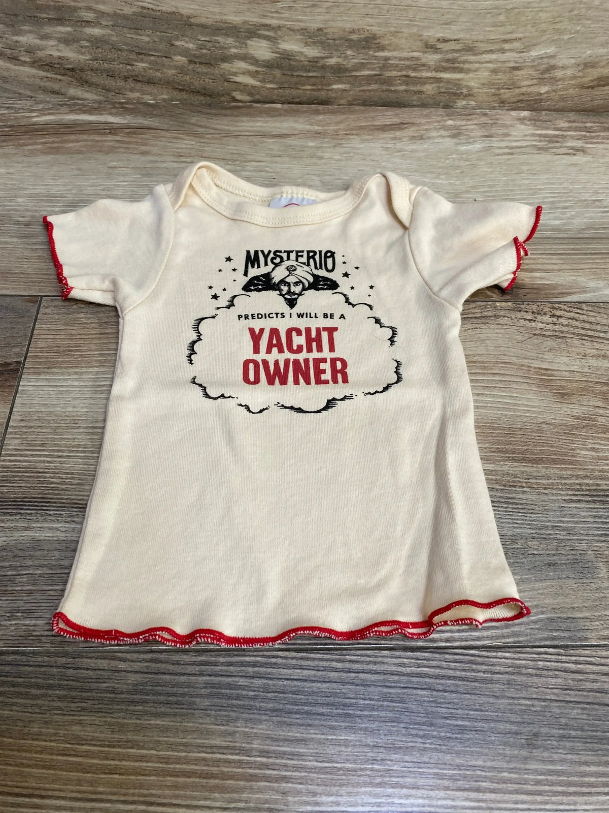 Flat-lay of cream newborn Wrybaby short-sleeve shirt with red lettuce-edge trim and front graphic YACHT OWNER on wood floor