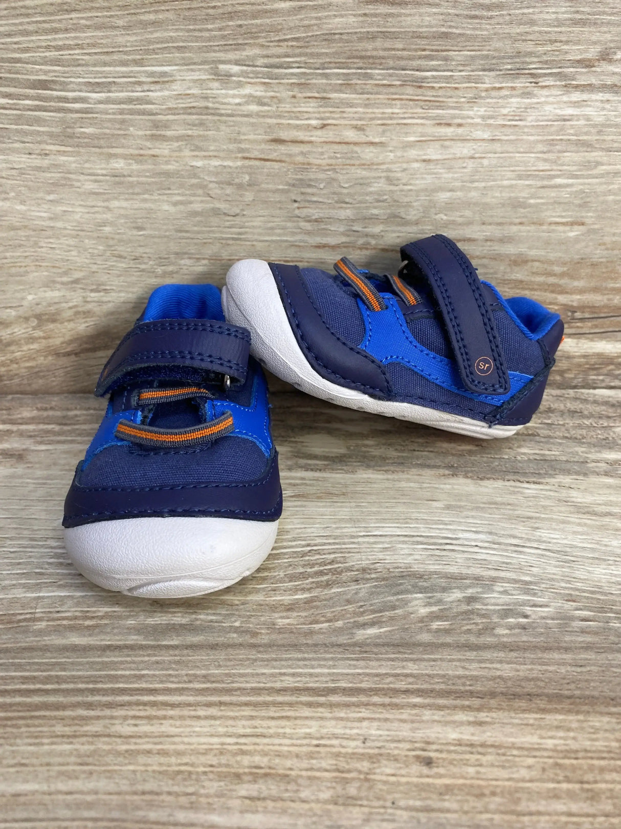 Front and side view of Stride Rite Kylin toddler sneakers showing navy uppers, white toe, orange accents, velcro strap on wood